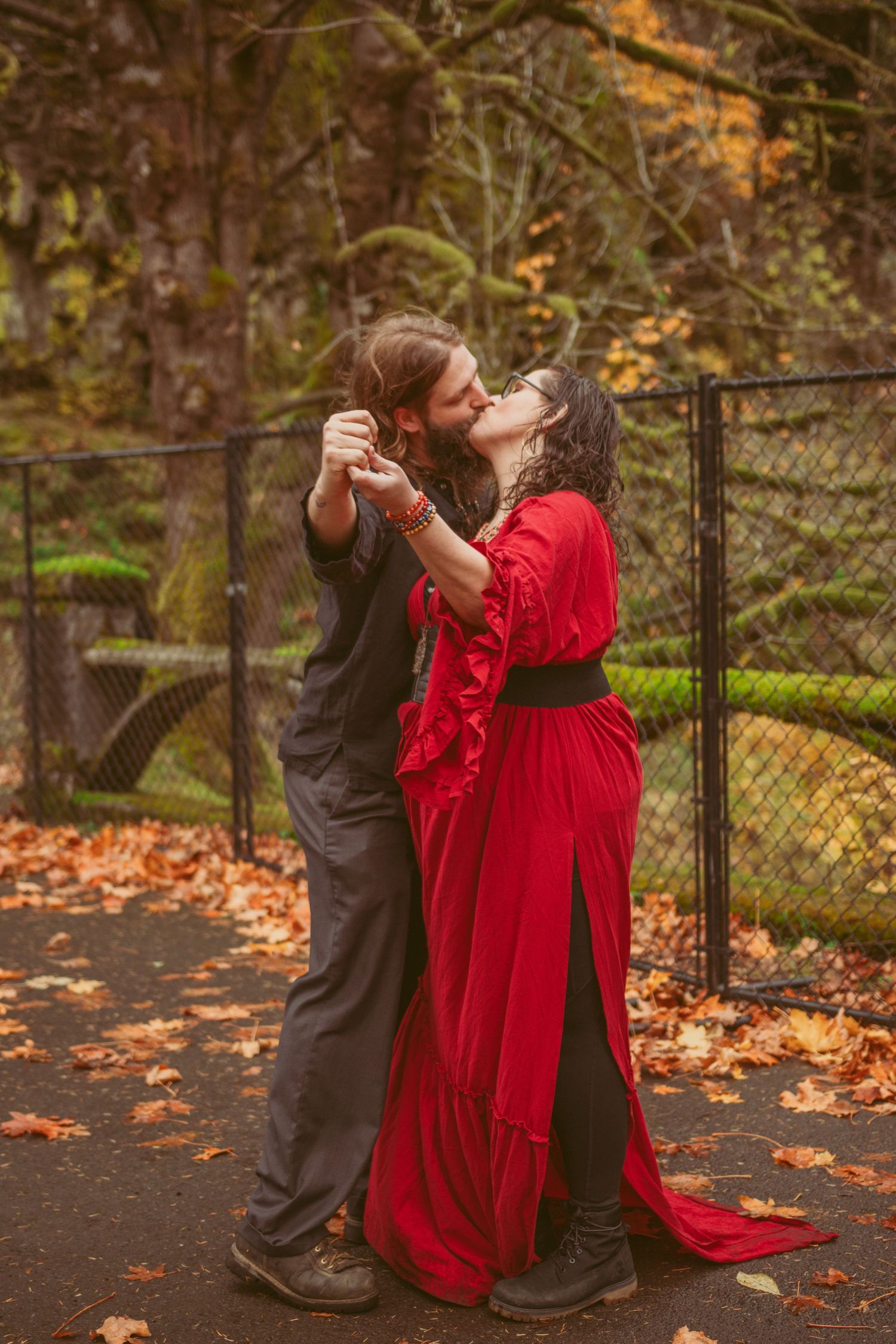 A couple in red and black attire dance together on fallen autumn leaves in a romantic outdoor setting.
