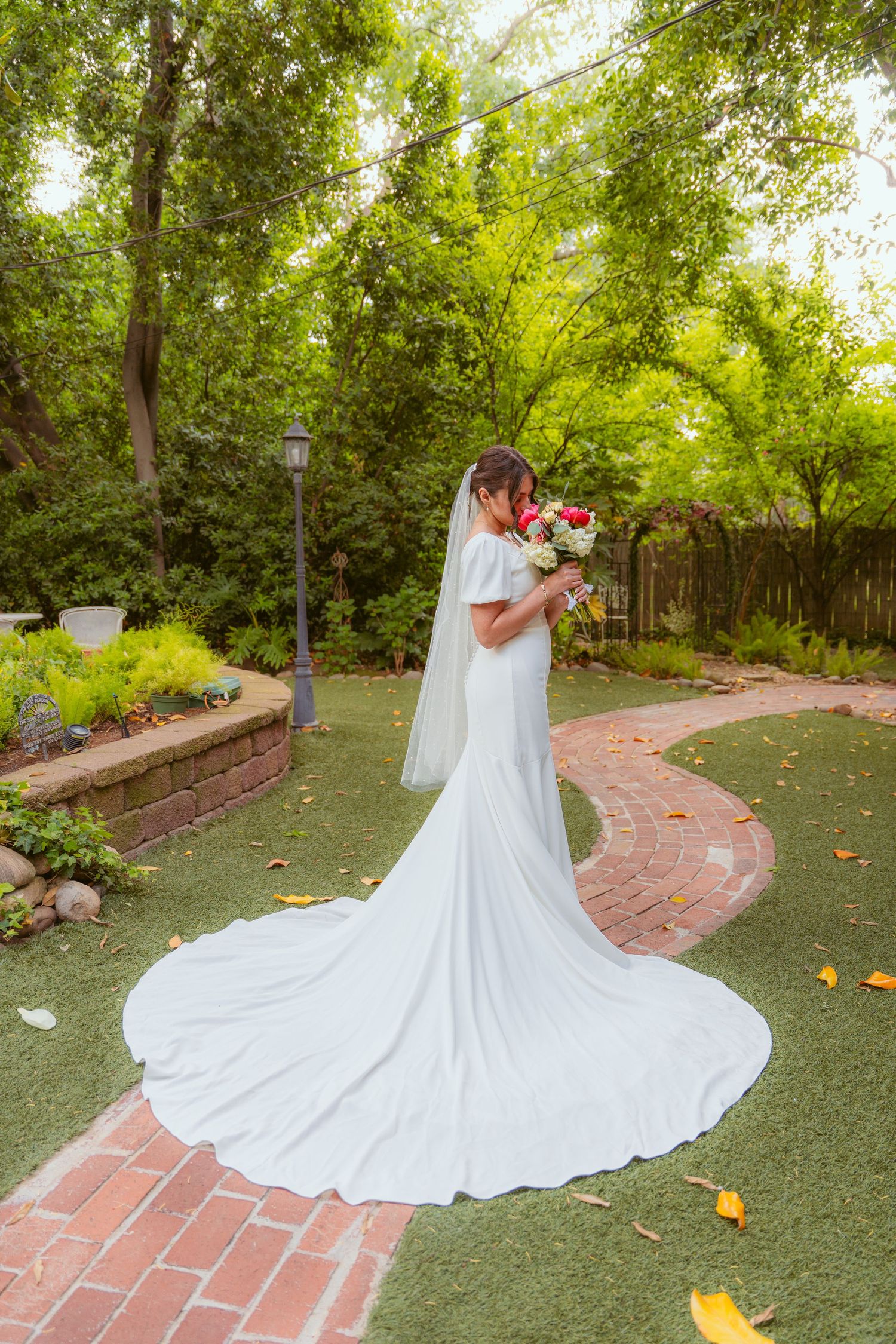 Flowing white wedding gown train extends across garden path surrounded by green trees and fallen leaves.