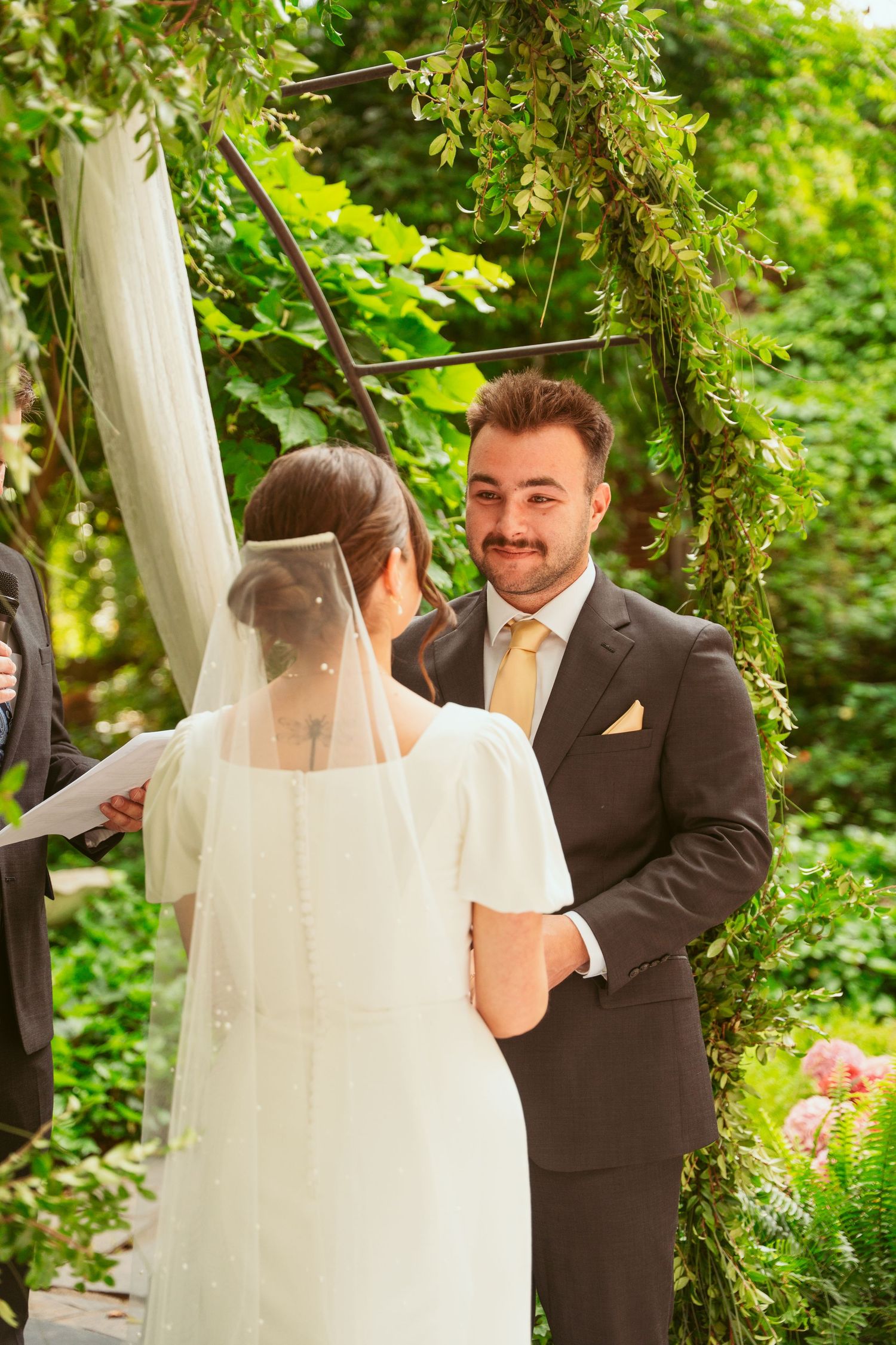 A couple shares their first moments as newlyweds under a garden arch decorated with white fabric.