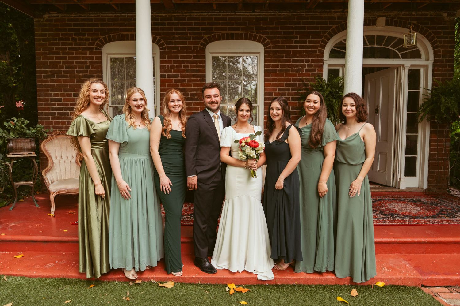 Wedding party in sage green and mint bridesmaid dresses poses on red carpet outside brick mansion.
