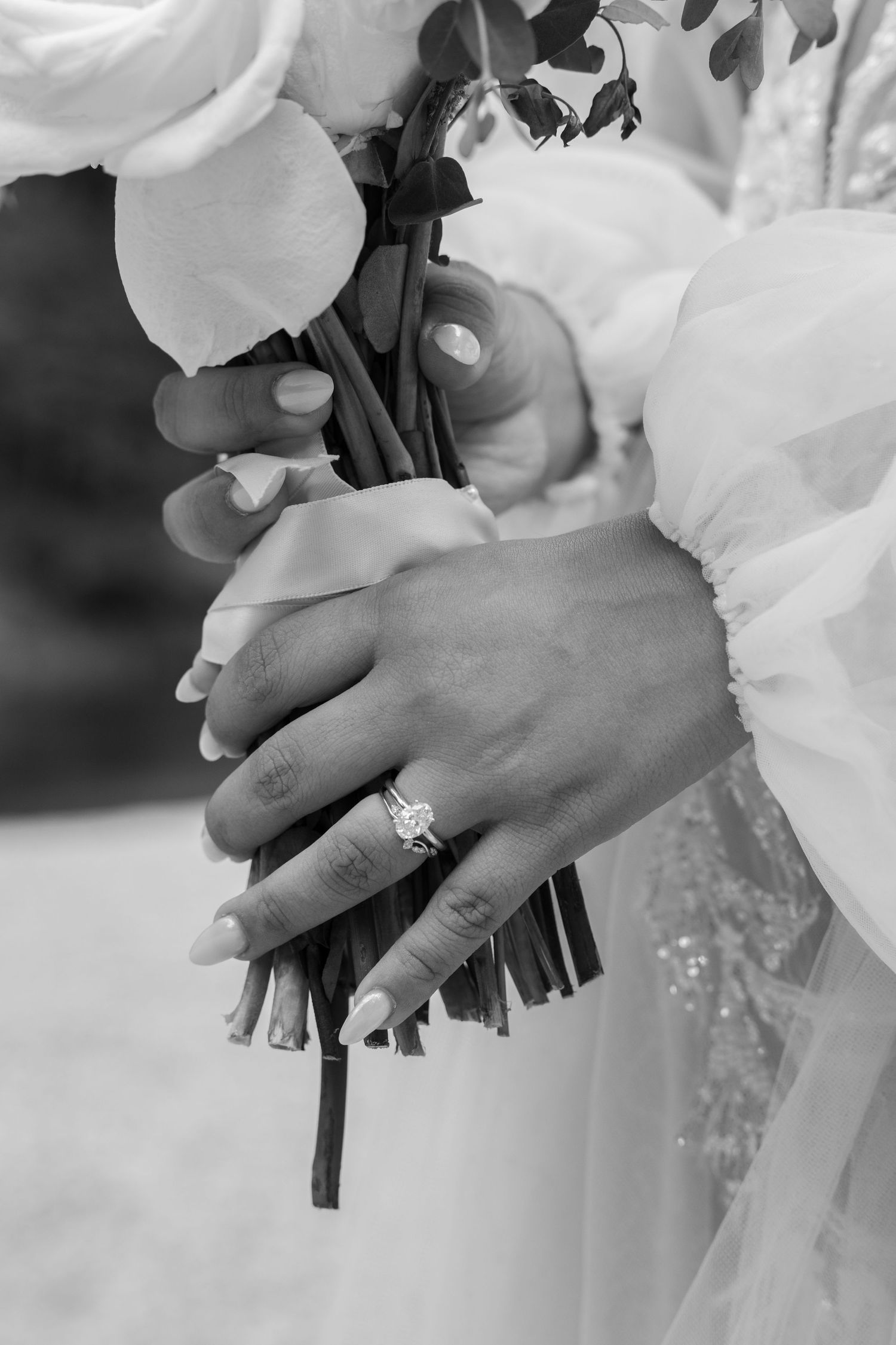 Black and white detail shot of hands holding bridal bouquet with engagement ring.