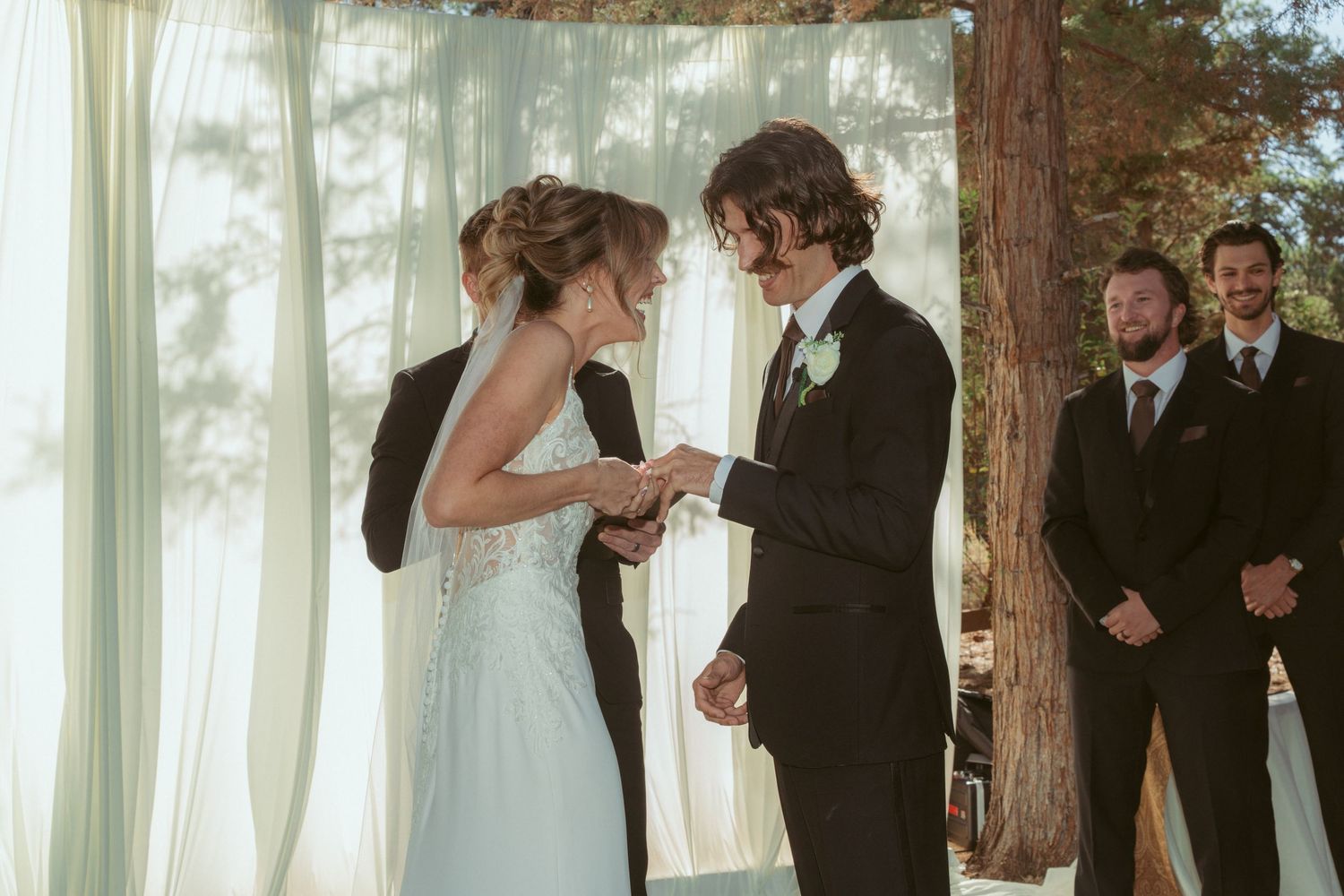 Wedding ceremony participants sharing joyful moments under tall trees with white drapes.