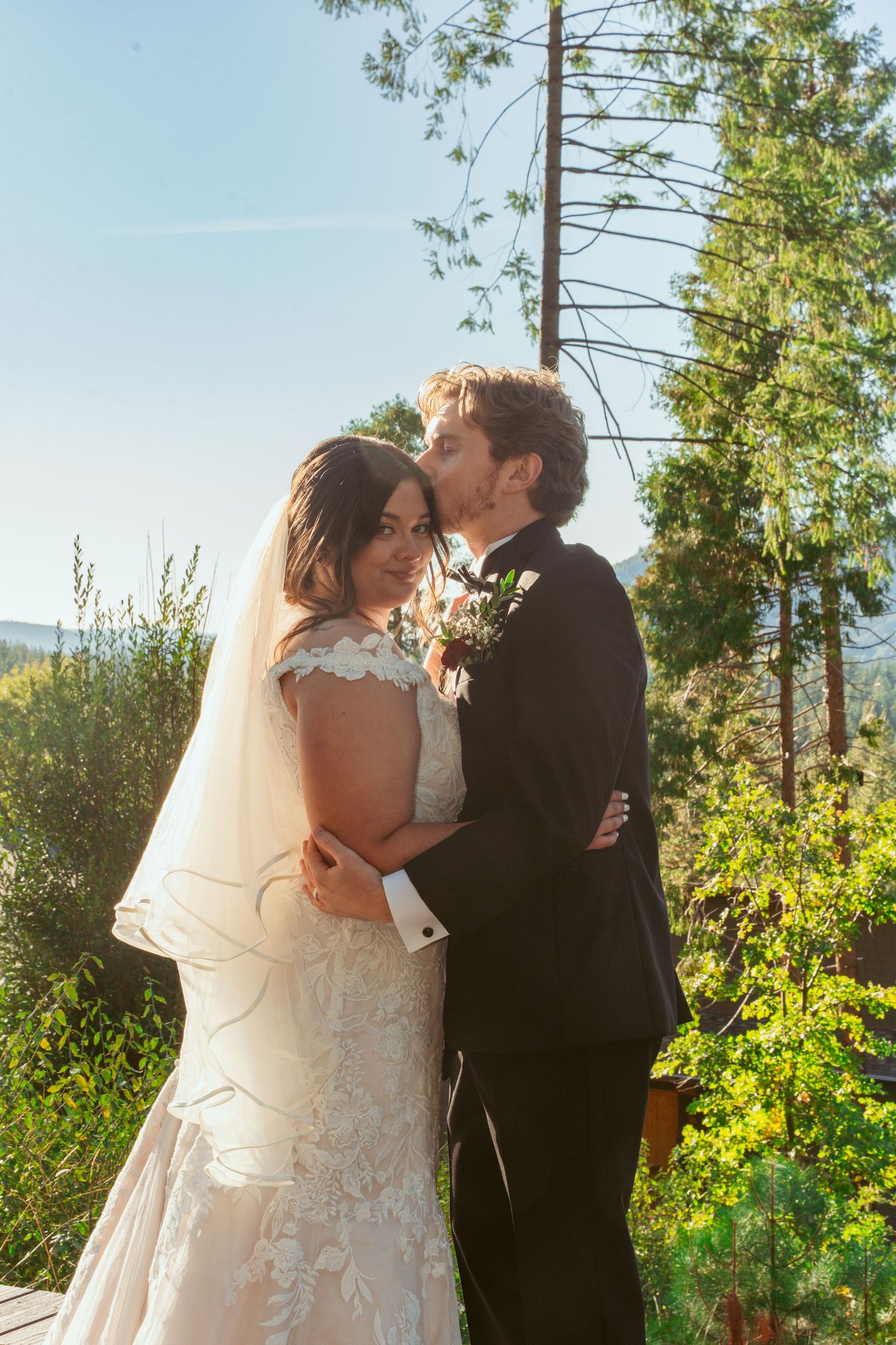 Tender moment between newlyweds against a backdrop of tall pine trees and blue sky.