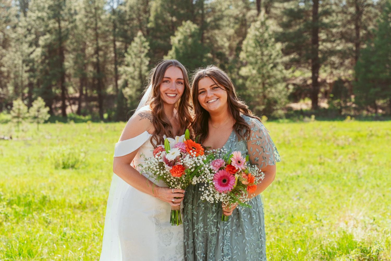 Wedding party members smile and laugh while holding coral flower bouquets in outdoor forest setting.