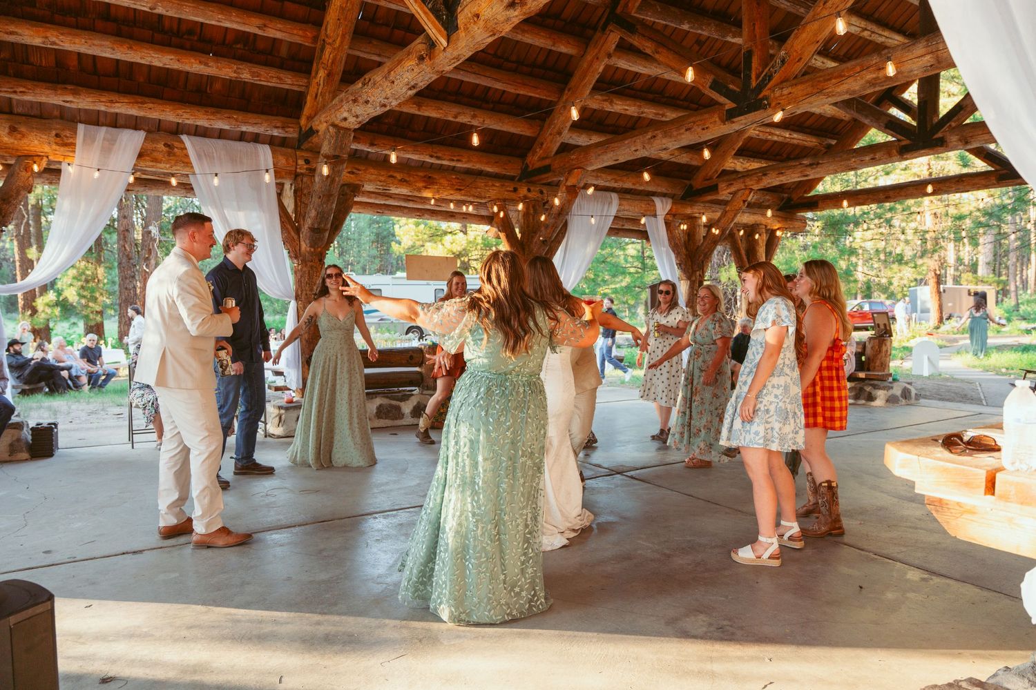 Wedding celebration with guests dancing under a rustic wooden pavilion in a forest setting.