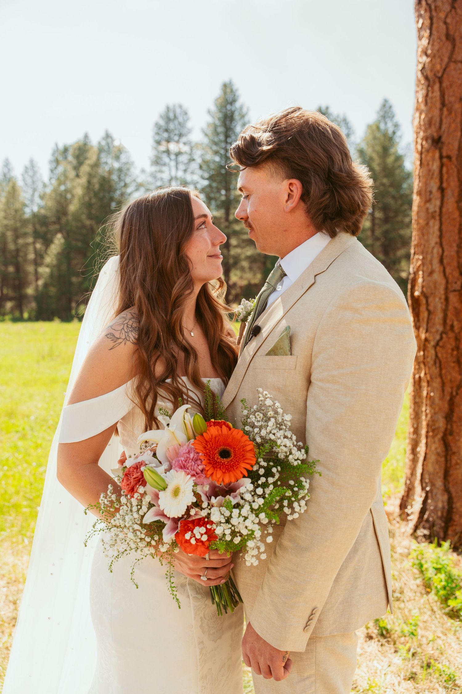 An intimate wedding moment captured beside a rustic pine tree with a colorful floral bouquet.