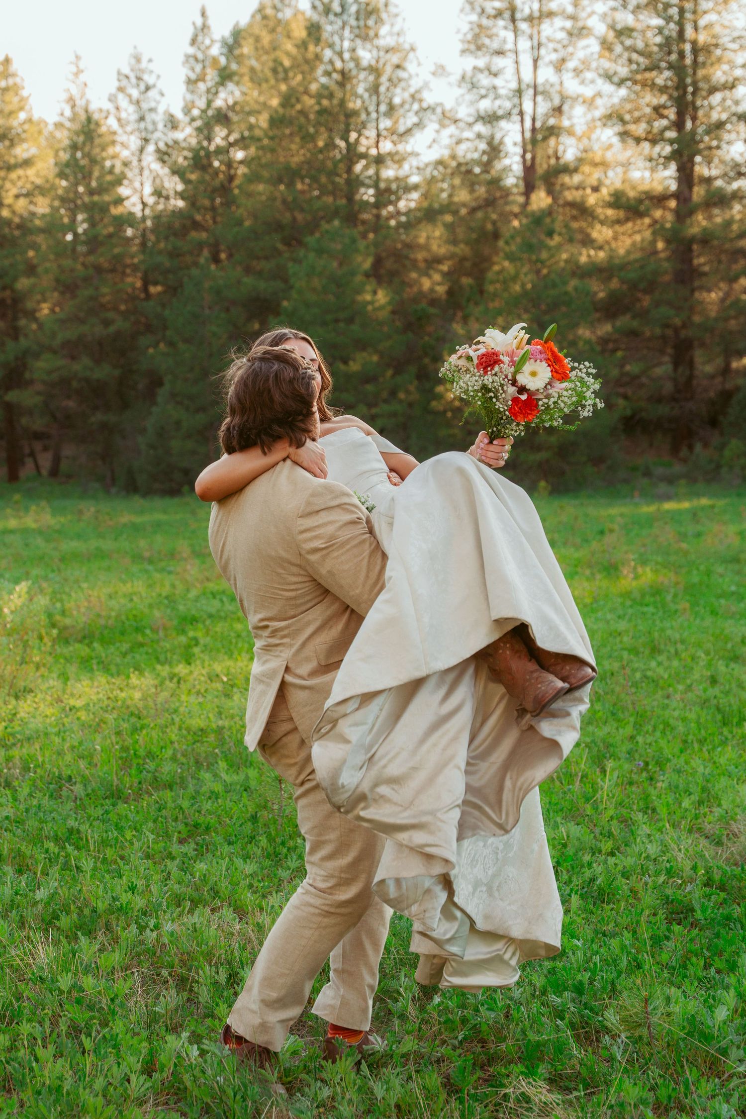 A playful wedding couple dances in a grassy meadow surrounded by tall pine trees at sunset.