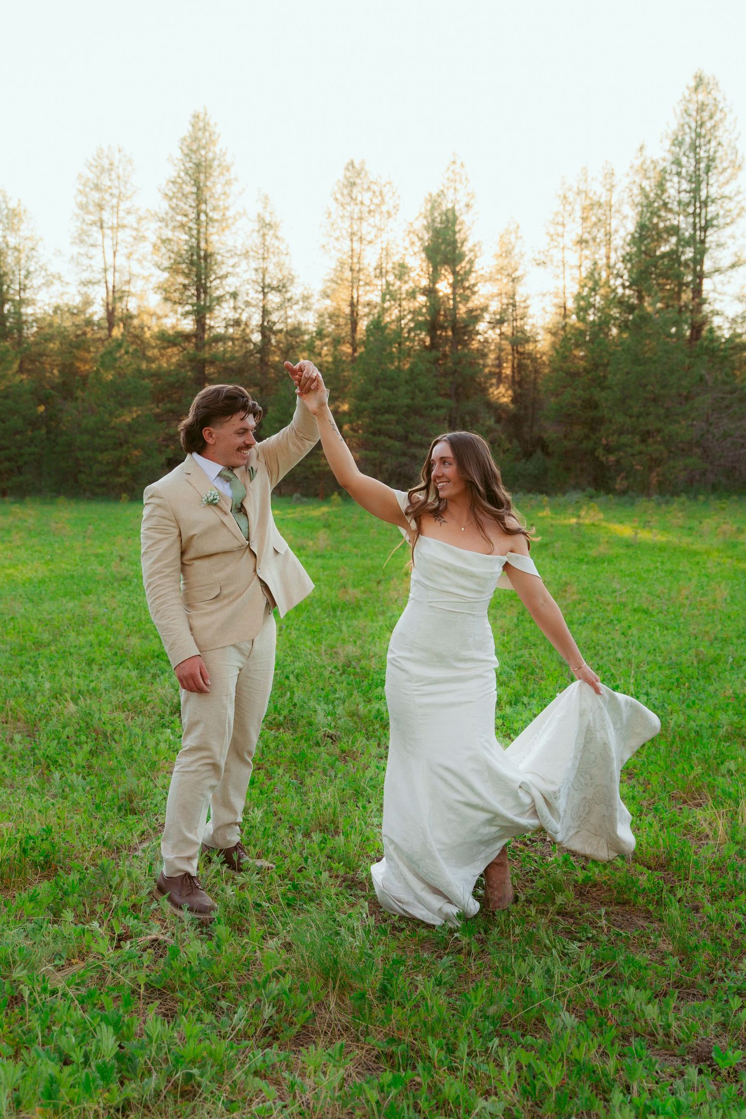 Couple in wedding attire dancing playfully in a meadow surrounded by evergreen trees.