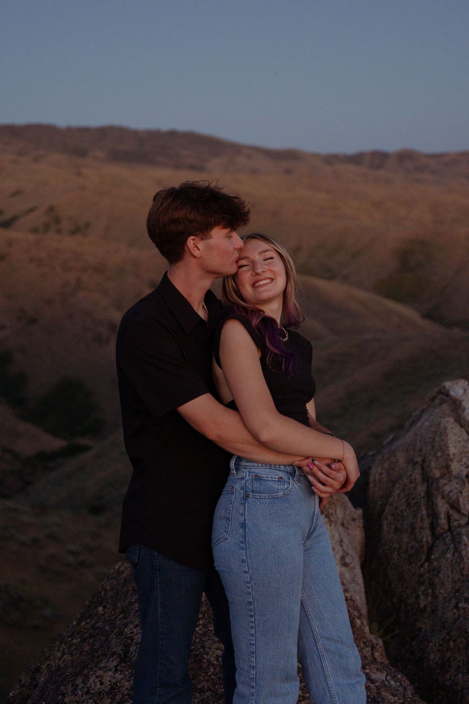 Couple sharing intimate moment against backdrop of rolling hills at sunset.