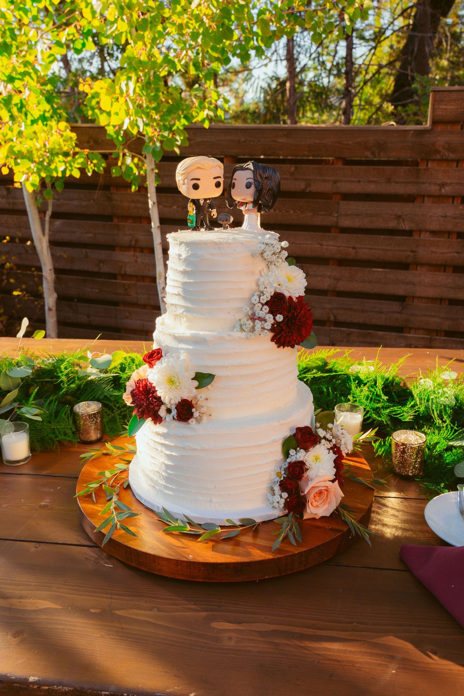 Three-tiered white wedding cake with red and pink floral decorations displayed on a wooden surface in natural lighting.