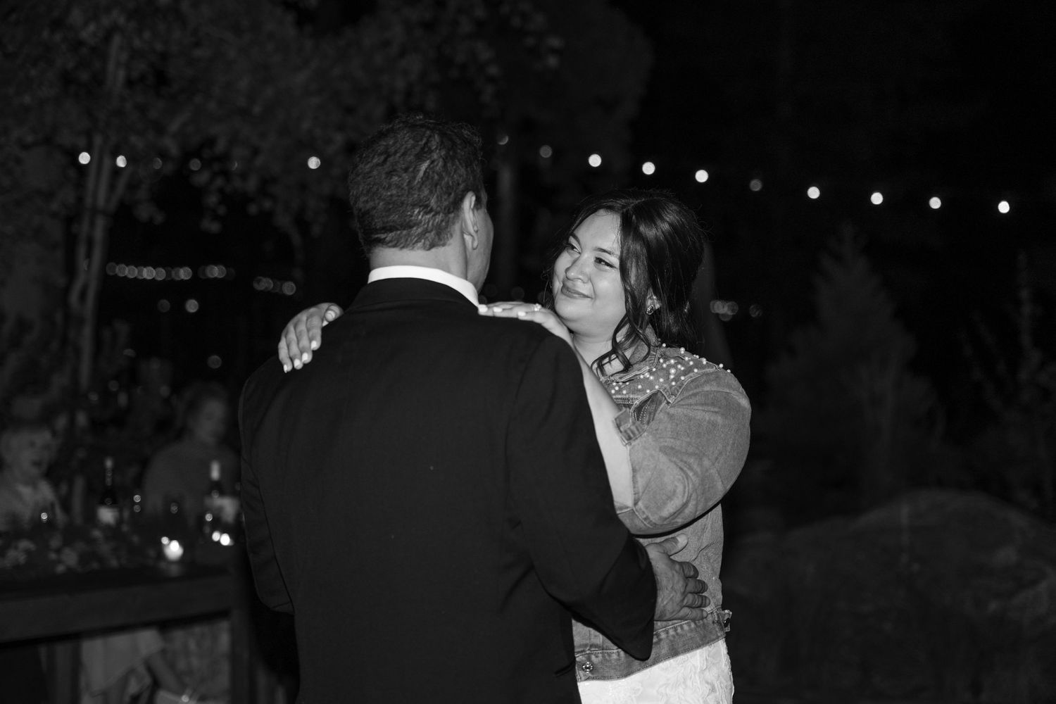 Dancing couples celebrate at a nighttime wedding reception under twinkling lights.