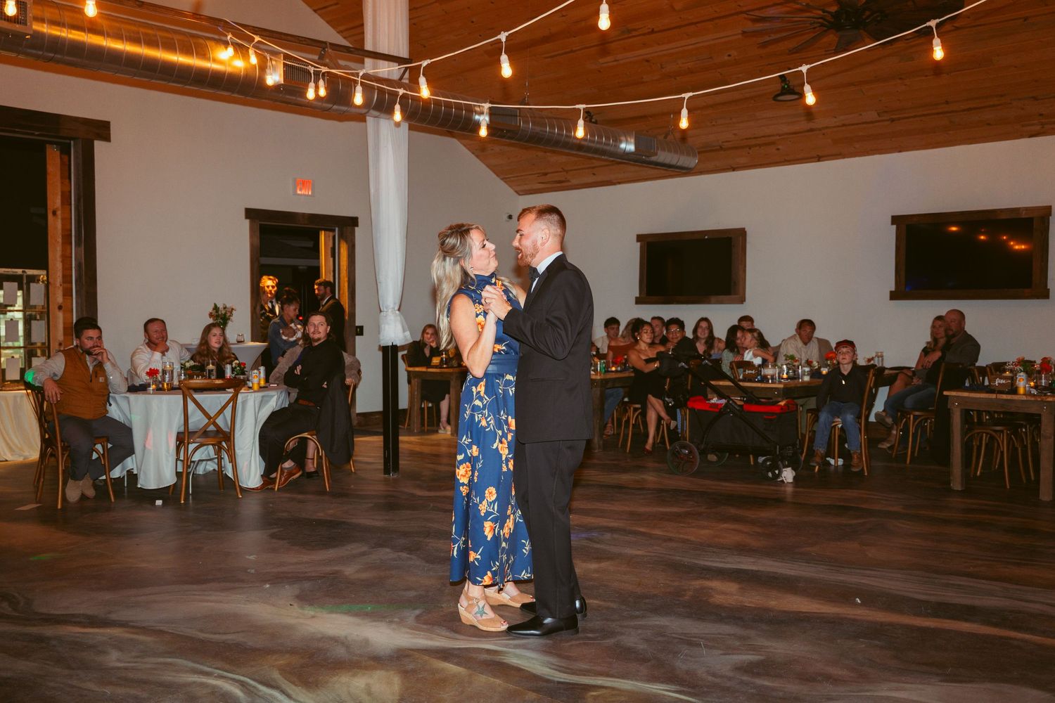 A couple dances in a rustic venue with string lights and wooden beams overhead.