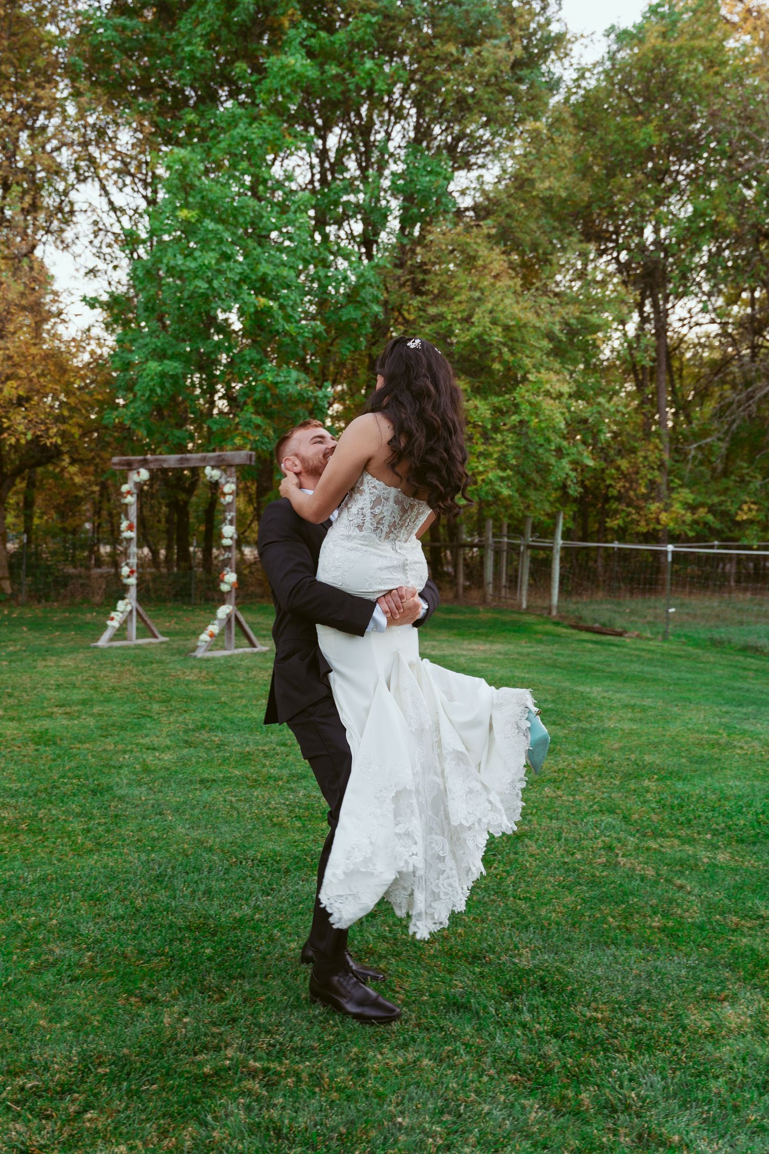 Playful wedding photo of couple dancing on grass with groom lifting bride in flowing white dress.