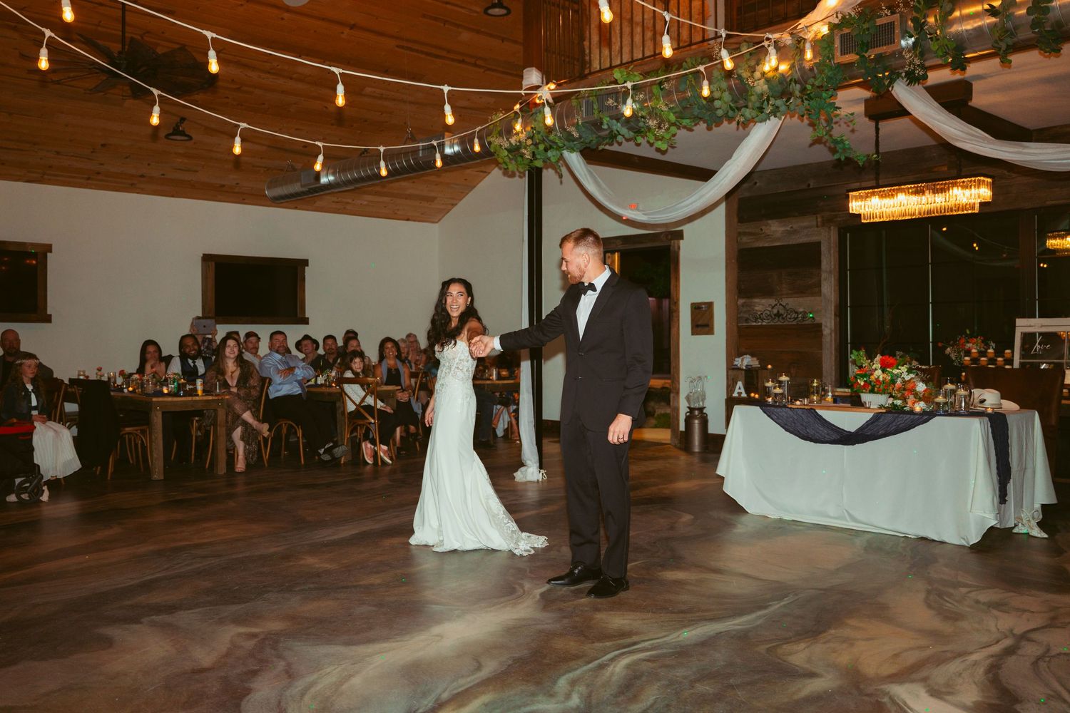 A couple performs their first dance at a wedding reception under string lights in a rustic venue.