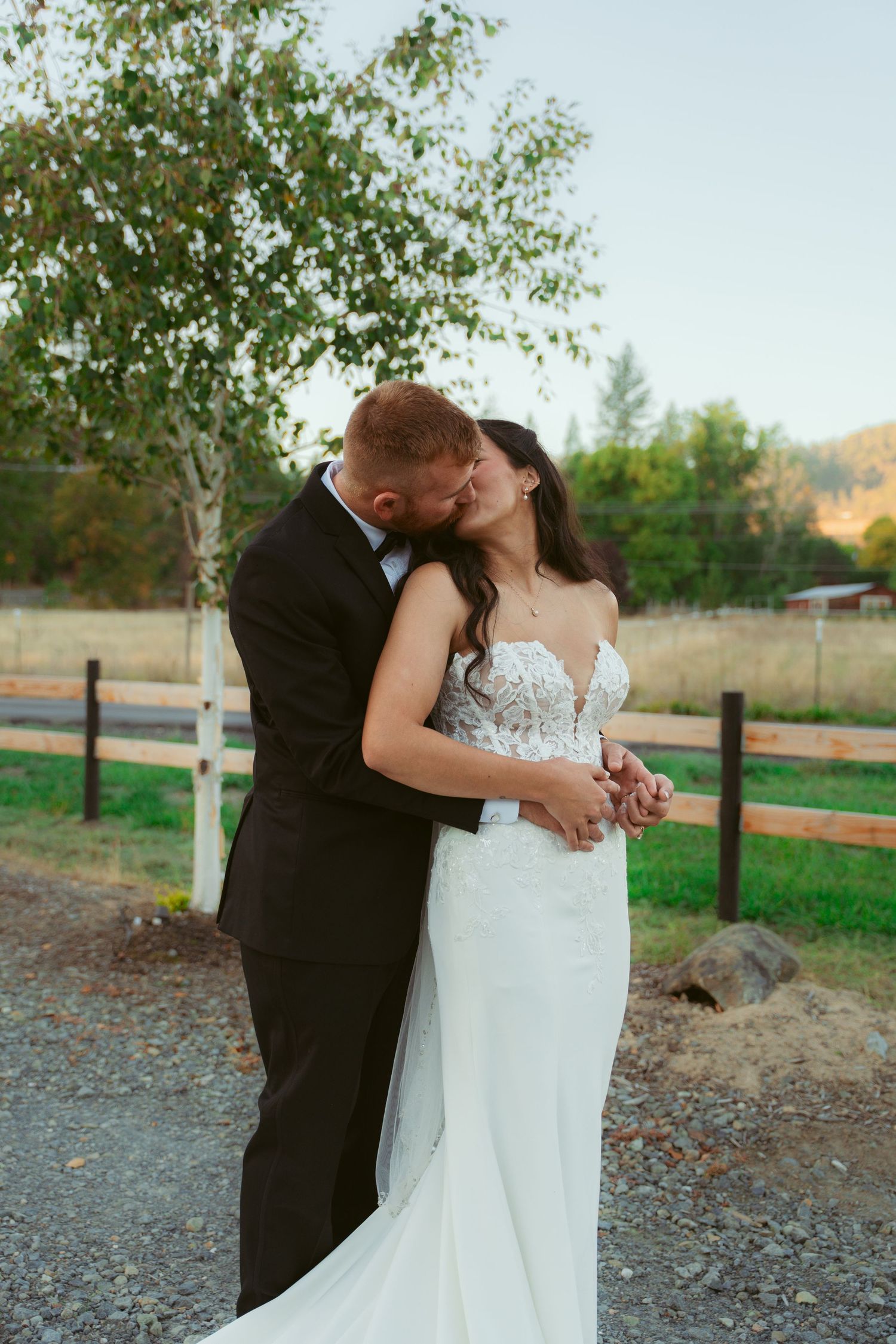 Romantic wedding couple embracing against a rustic fence and countryside backdrop.
