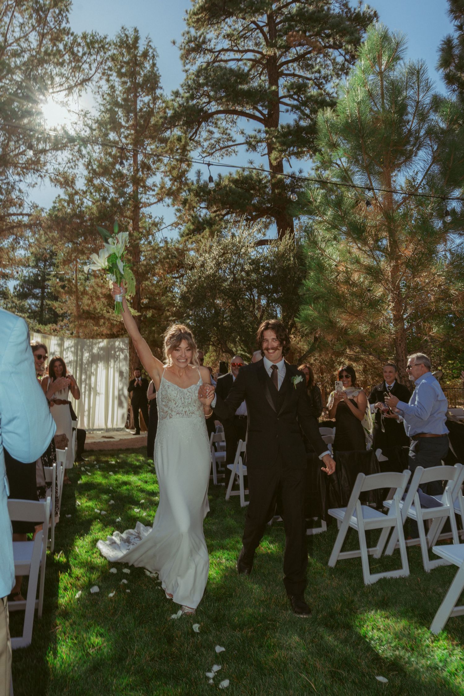 Wedding ceremony setup in a pine forest with white chairs and guests gathering on a grassy area.