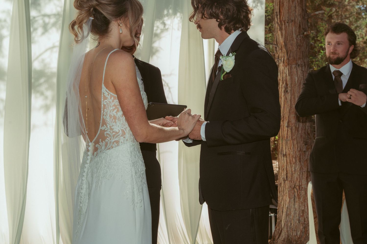 A couple shares their wedding vows beneath tall trees and white curtains at an outdoor ceremony.