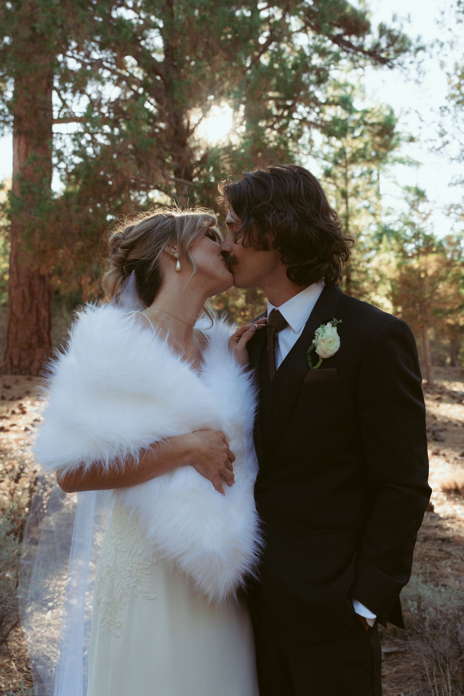 A romantic wedding couple shares a tender kiss in a pine forest setting while wearing winter wedding attire.