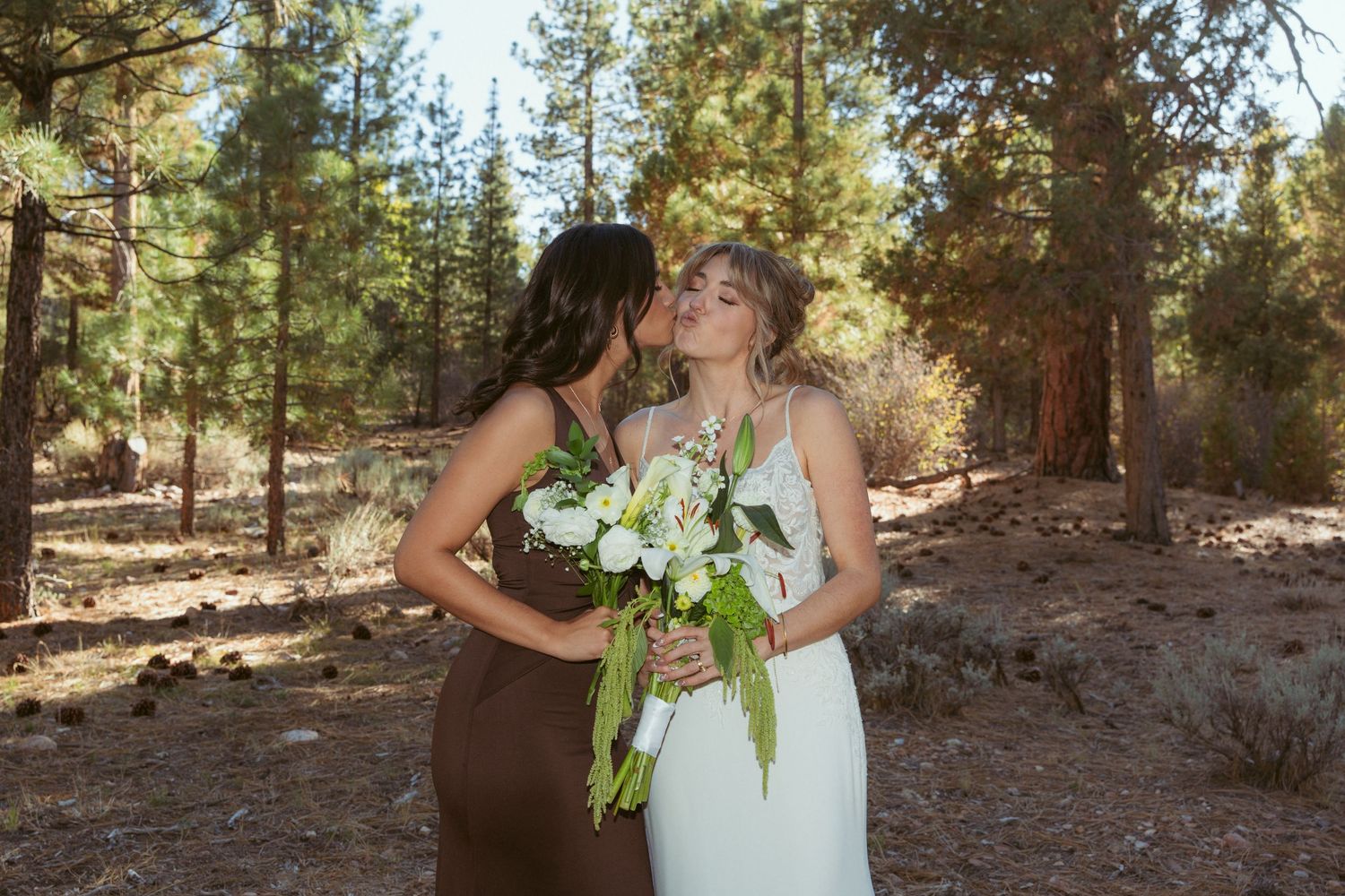 Two people embrace while holding white bouquets against a backdrop of tall pine trees.