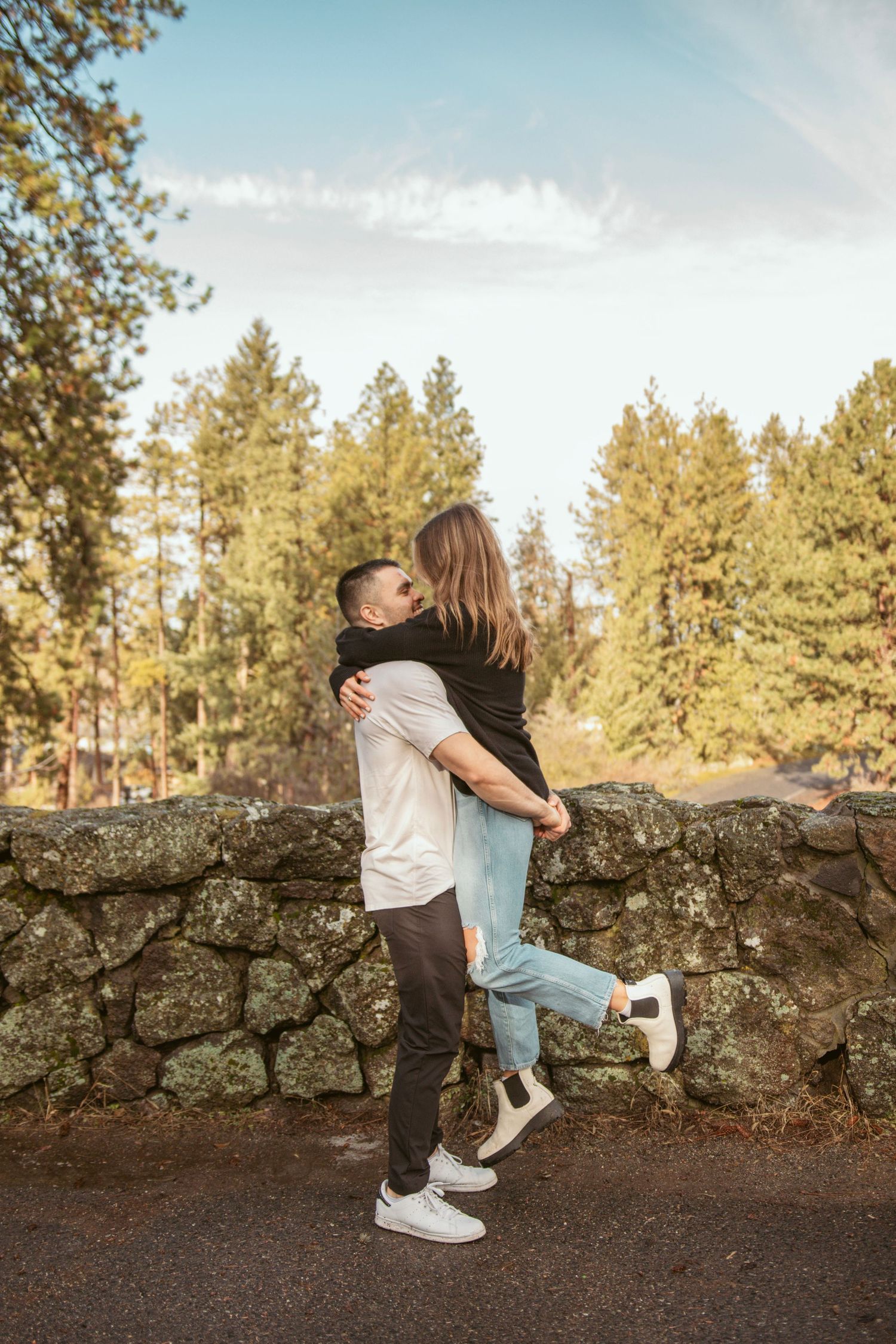 Playful couple poses against stone wall with evergreen forest backdrop.