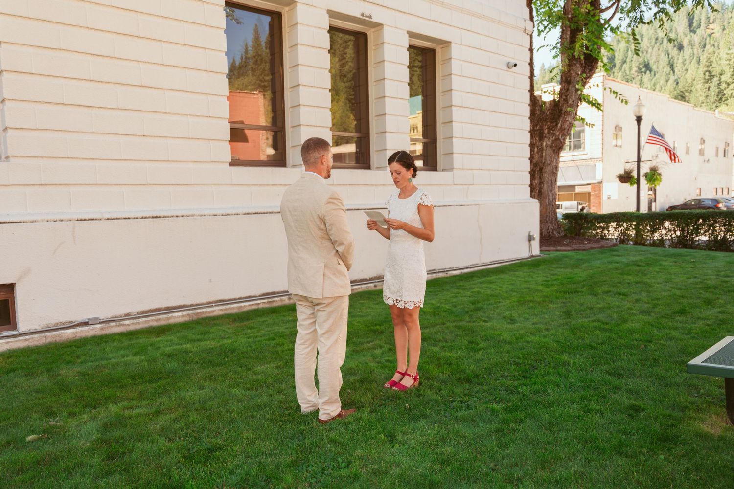 A couple shares a private moment on a grassy lawn outside a white building during a wedding ceremony.