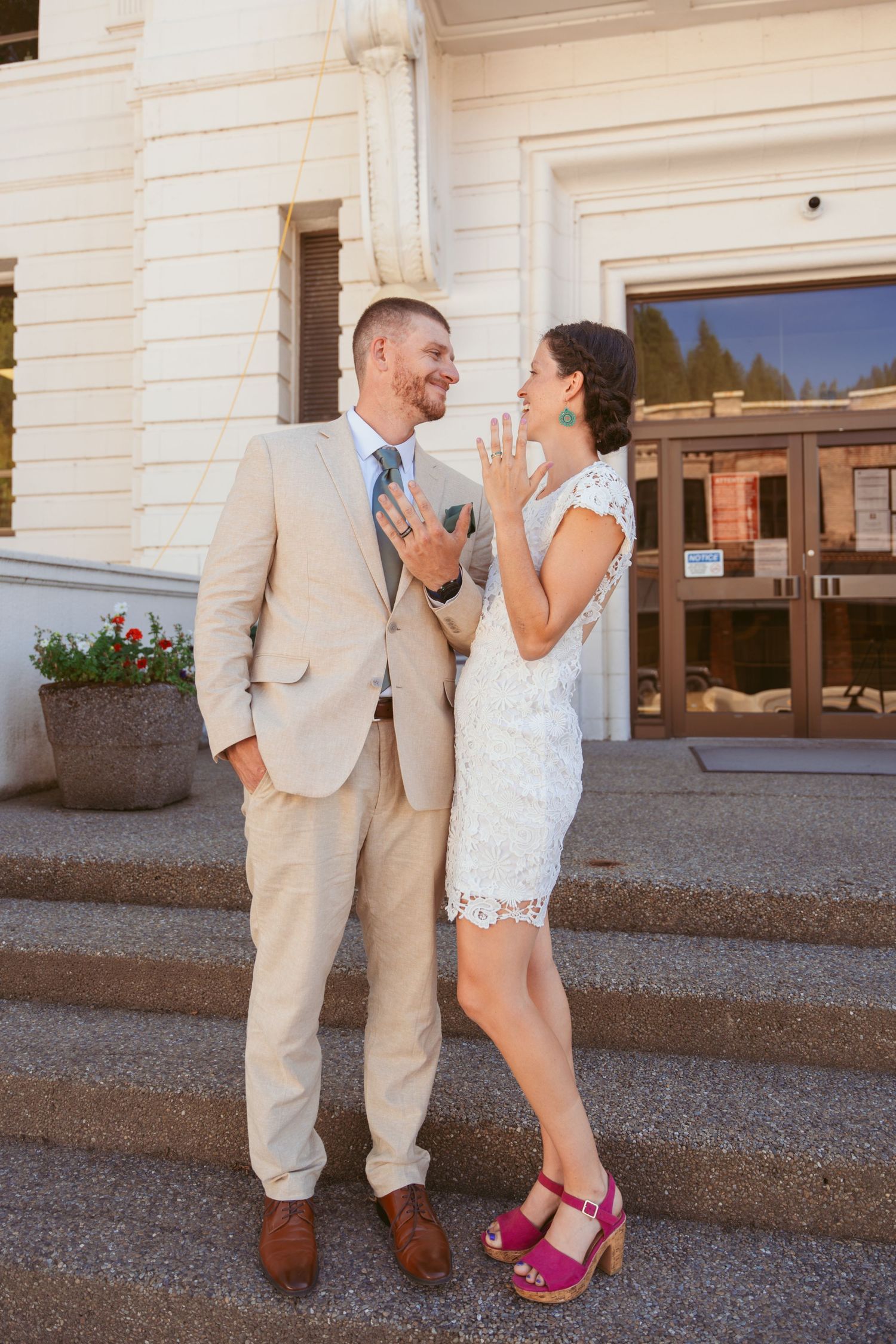Playful couple in wedding attire posing against white building exterior.