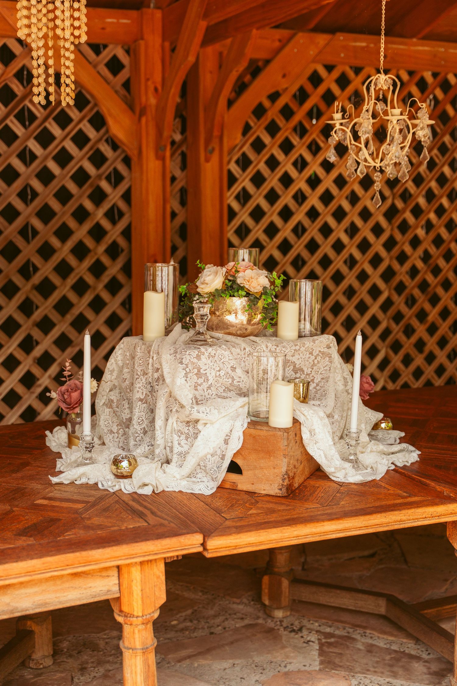 Elegant table setting with white lace, candles and floral arrangements under wooden lattice pergola.
