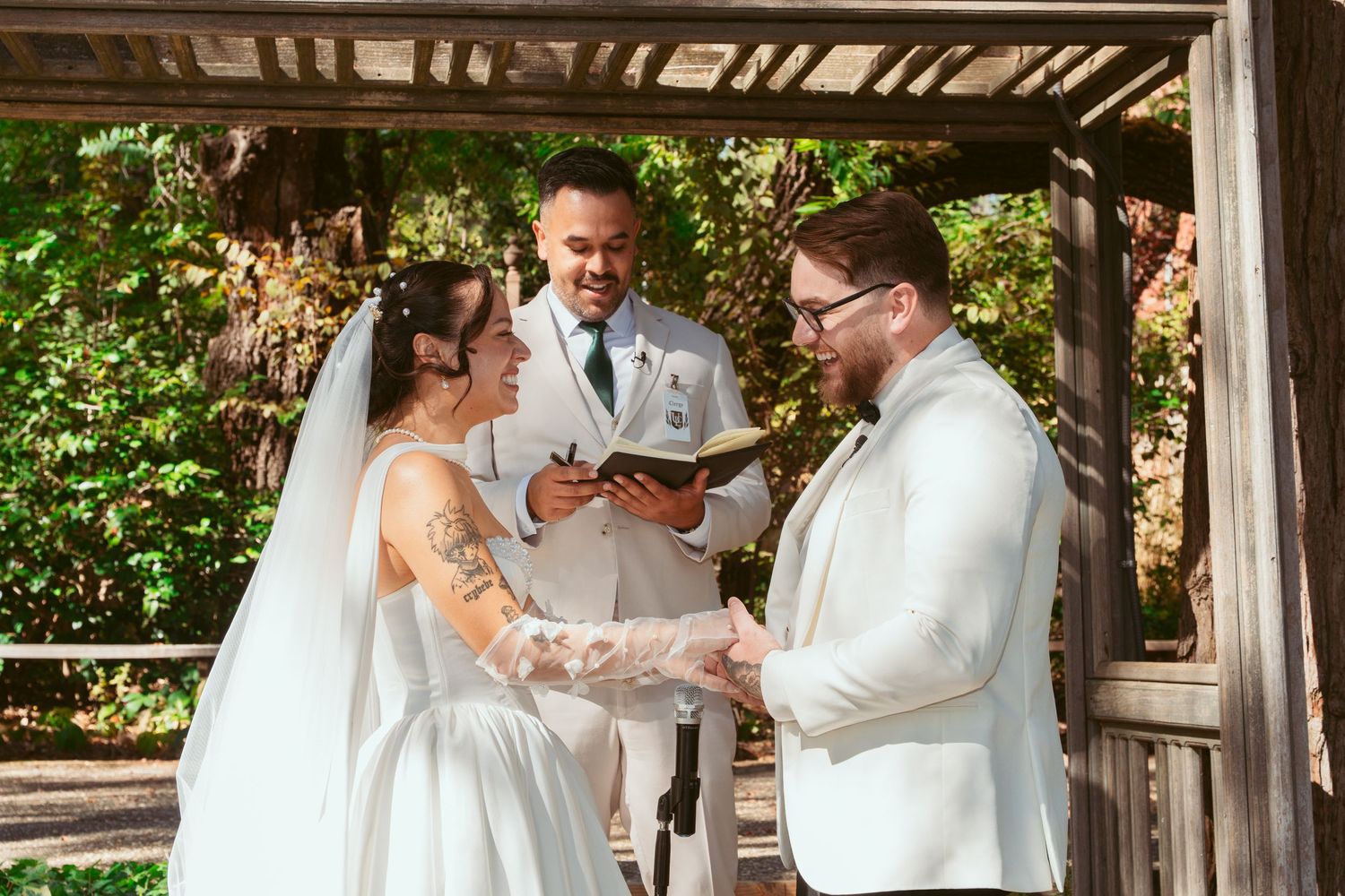 Wedding ceremony in progress under wooden pergola with officiant in white.