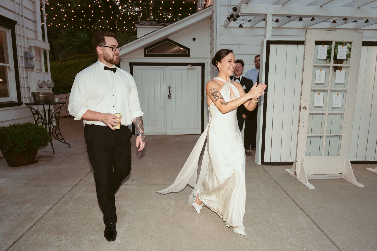 A couple walks along a concrete pathway outside a white building with string lights.