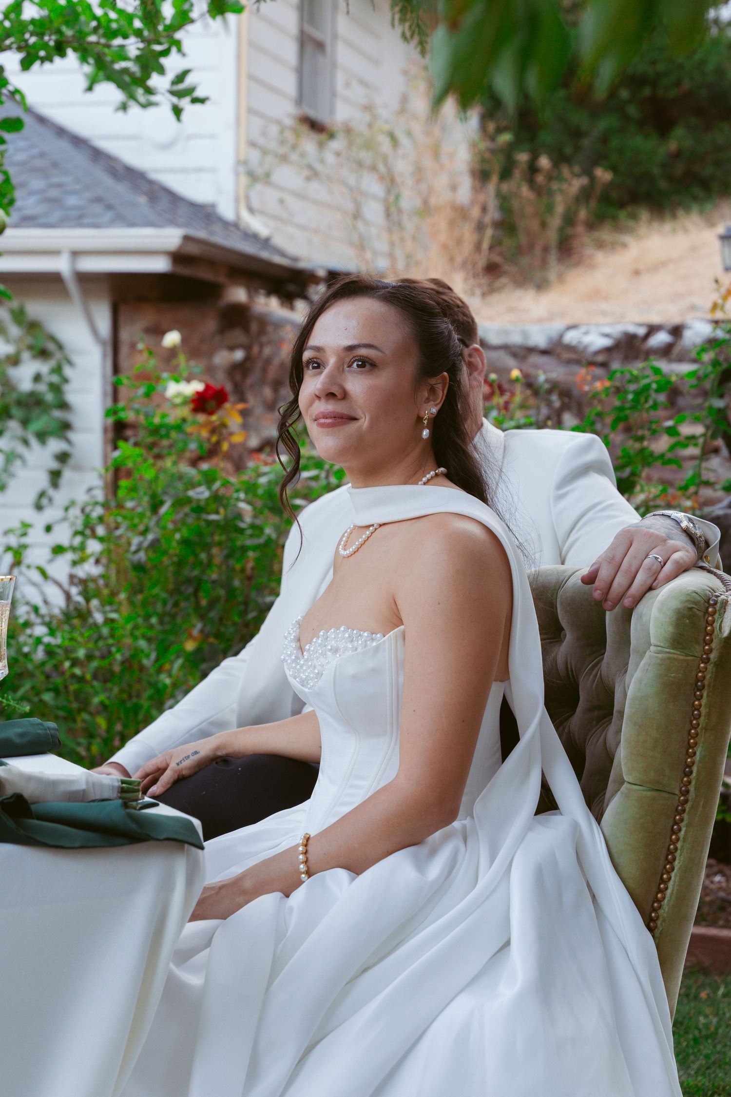 A bride in a white wedding gown sits elegantly on a vintage chair in a garden setting.