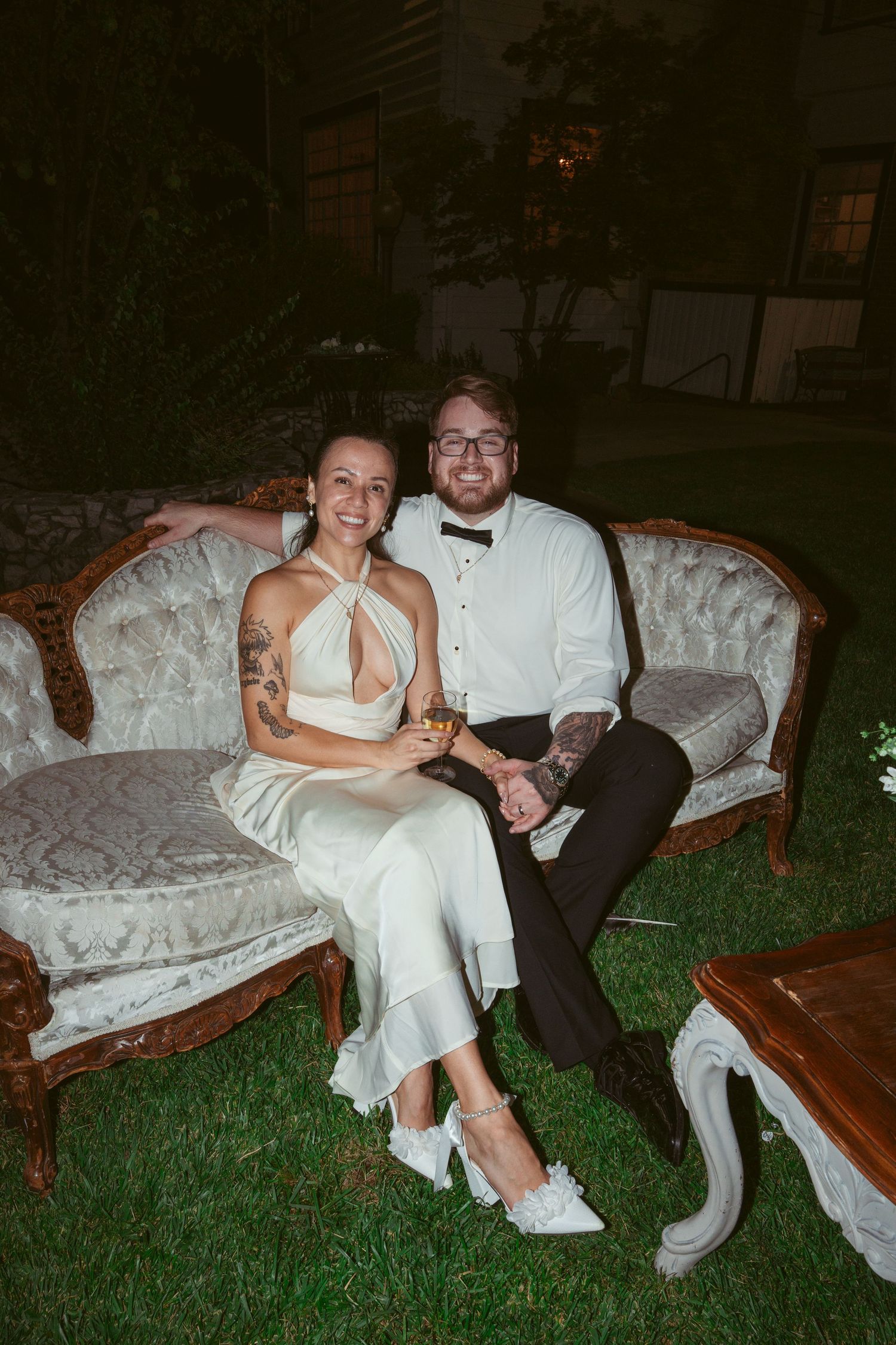 A couple relaxes on an elegant white couch placed outdoors on grass during an evening celebration.