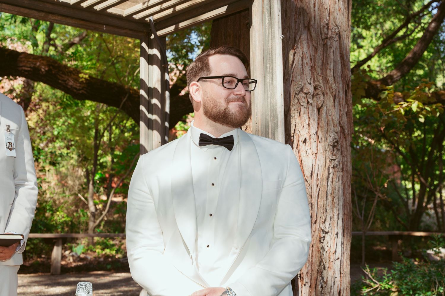 Wedding party stands in line under wooden pergola surrounded by lush greenery.