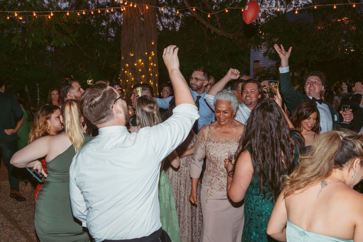 Wedding party dancing and celebrating under warm string lights at an outdoor reception.