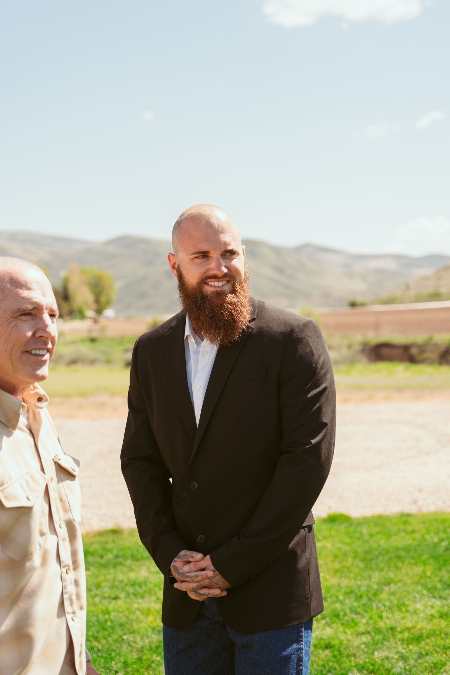 People in formal attire share laughs on a grassy field with mountains in the background.