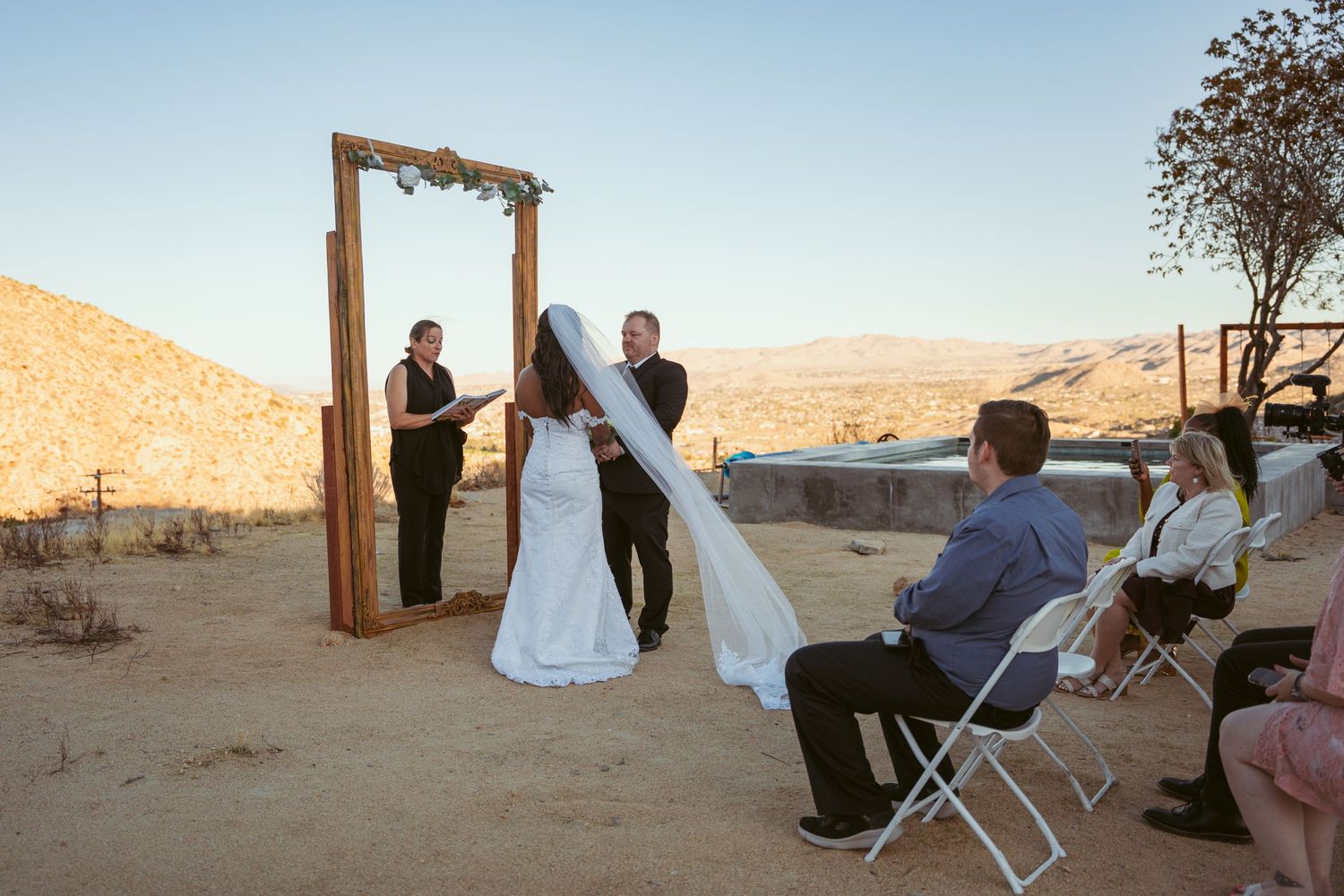 Small outdoor wedding ceremony with wooden arch and folding chairs in desert setting.