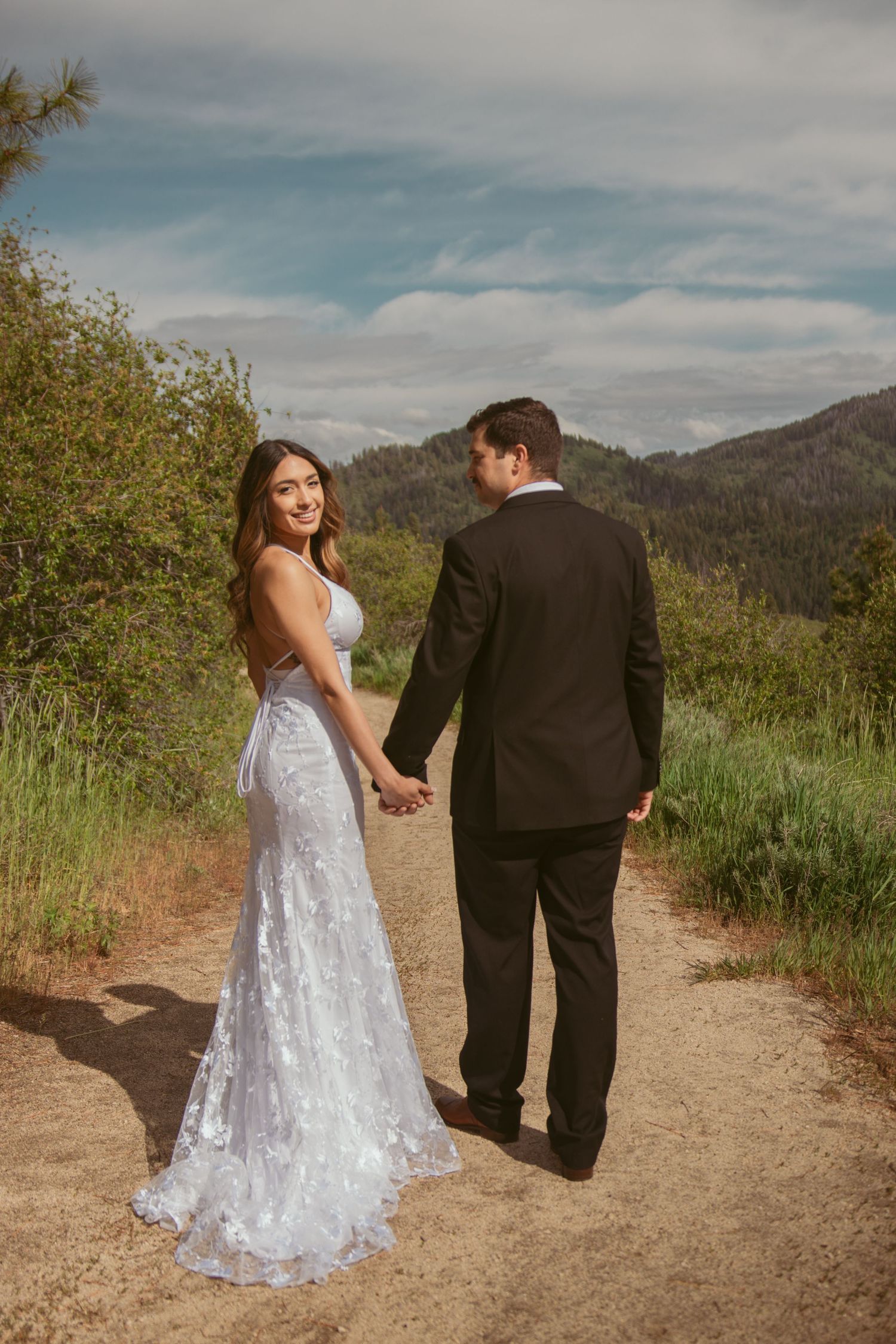 A couple walks hand in hand on a mountain trail in wedding attire.