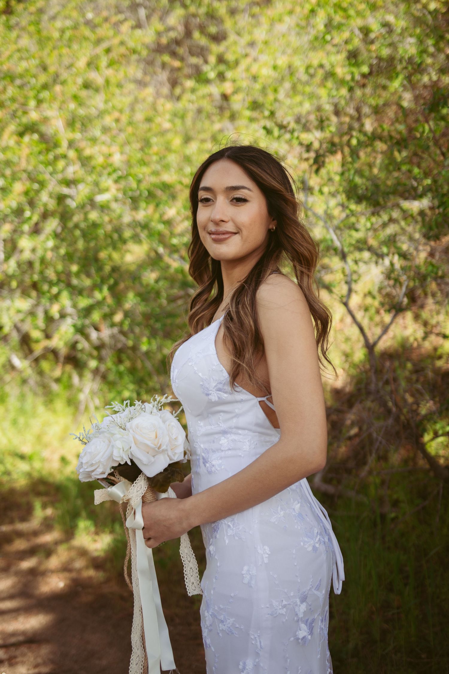 A bride in a white lace dress poses outdoors holding a bouquet against a green natural backdrop.