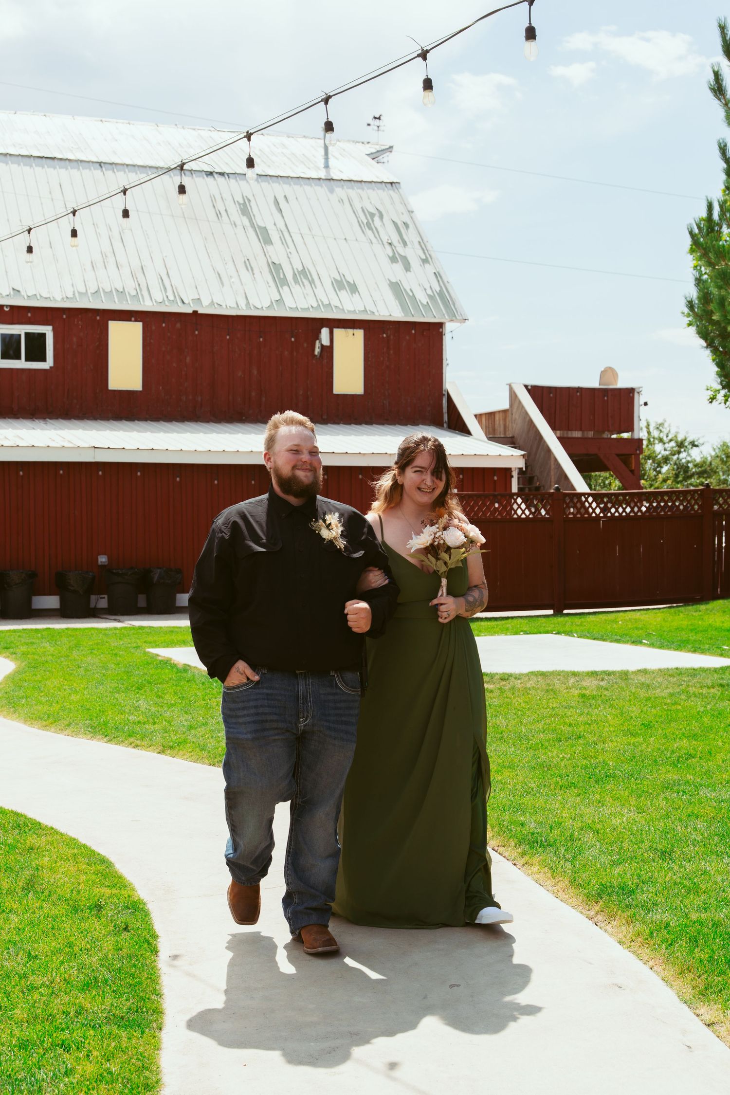 A couple walks along a concrete path next to a red barn on a sunny day, with string lights overhead.
