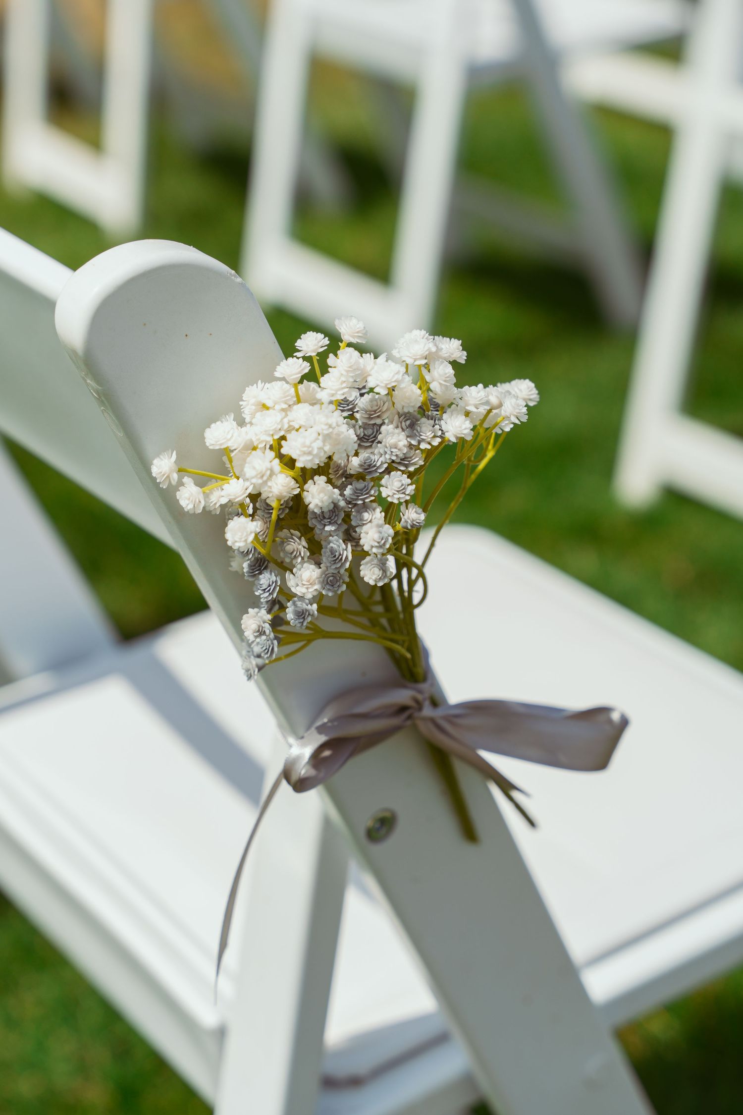 A delicate baby's breath flower arrangement tied with a gray ribbon decorates a white folding chair at an outdoor wedding.