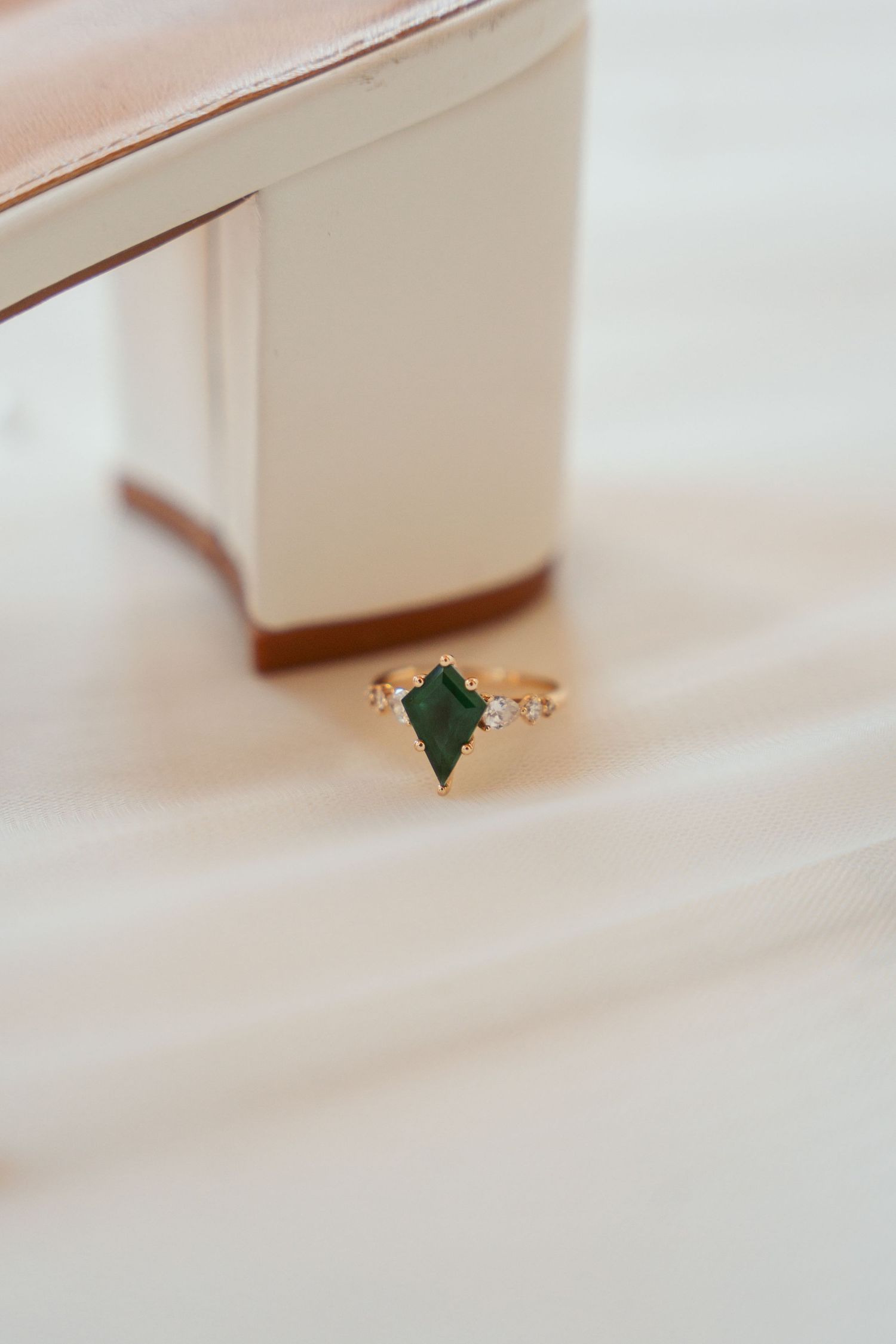 A delicate emerald engagement ring with diamond accents rests against a soft pink surface.