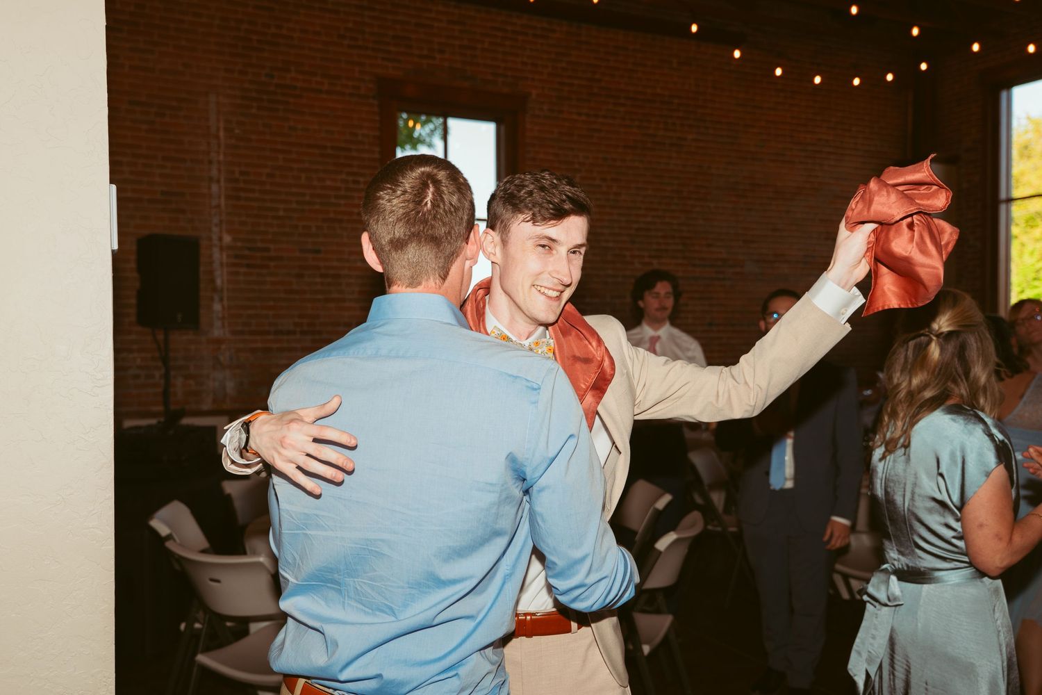 Wedding guests dance energetically in a rustic brick venue decorated with string lights.