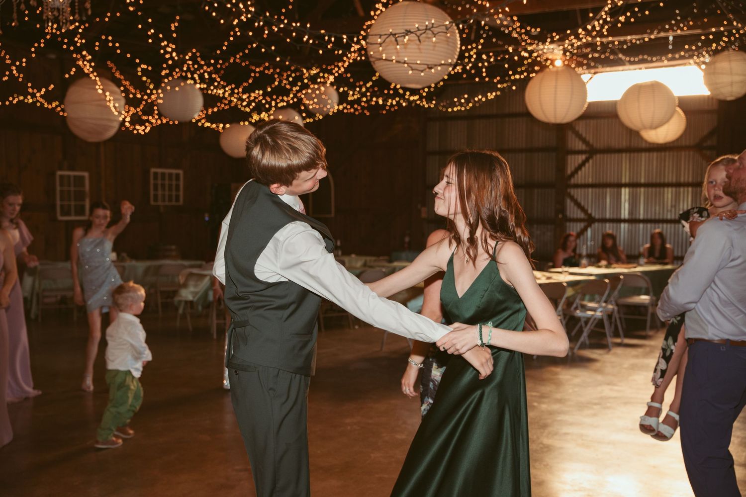 Wedding guests dancing energetically under warm string lights and paper lanterns at an indoor reception venue.