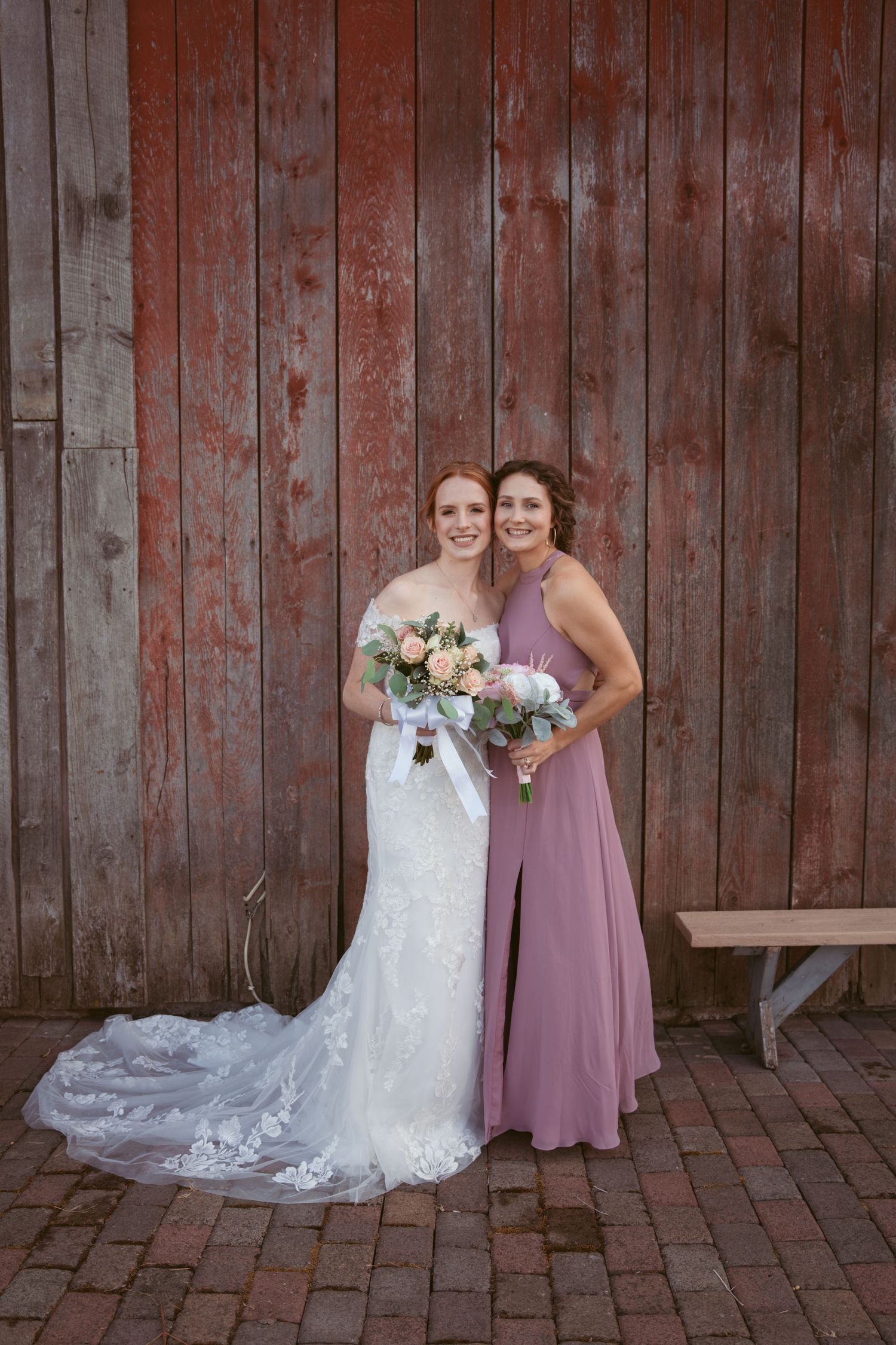 Wedding party poses in front of rustic red barn wall wearing mauve dresses and white lace gown.
