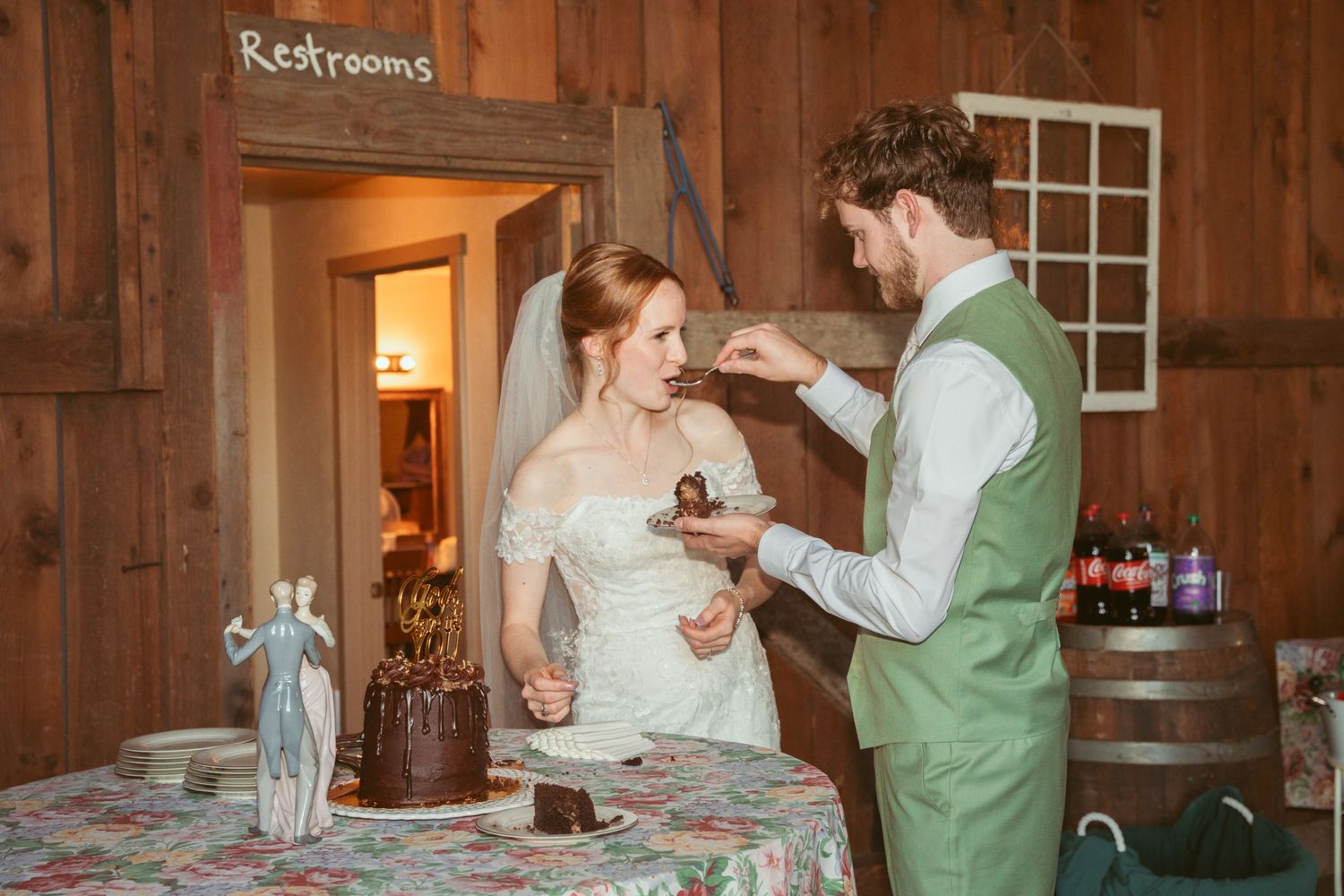 Bride and groom share wedding cake during their reception in a rustic barn setting.