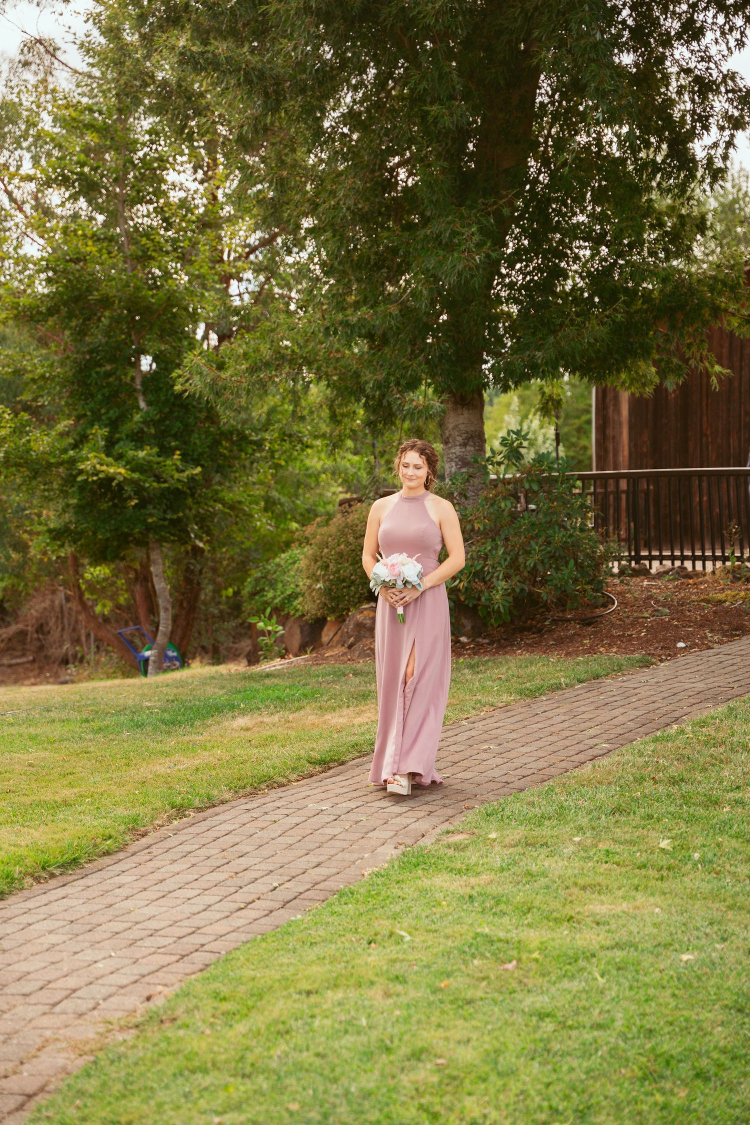 A person in a pink dress walks down a curved brick path surrounded by green grass and trees.
