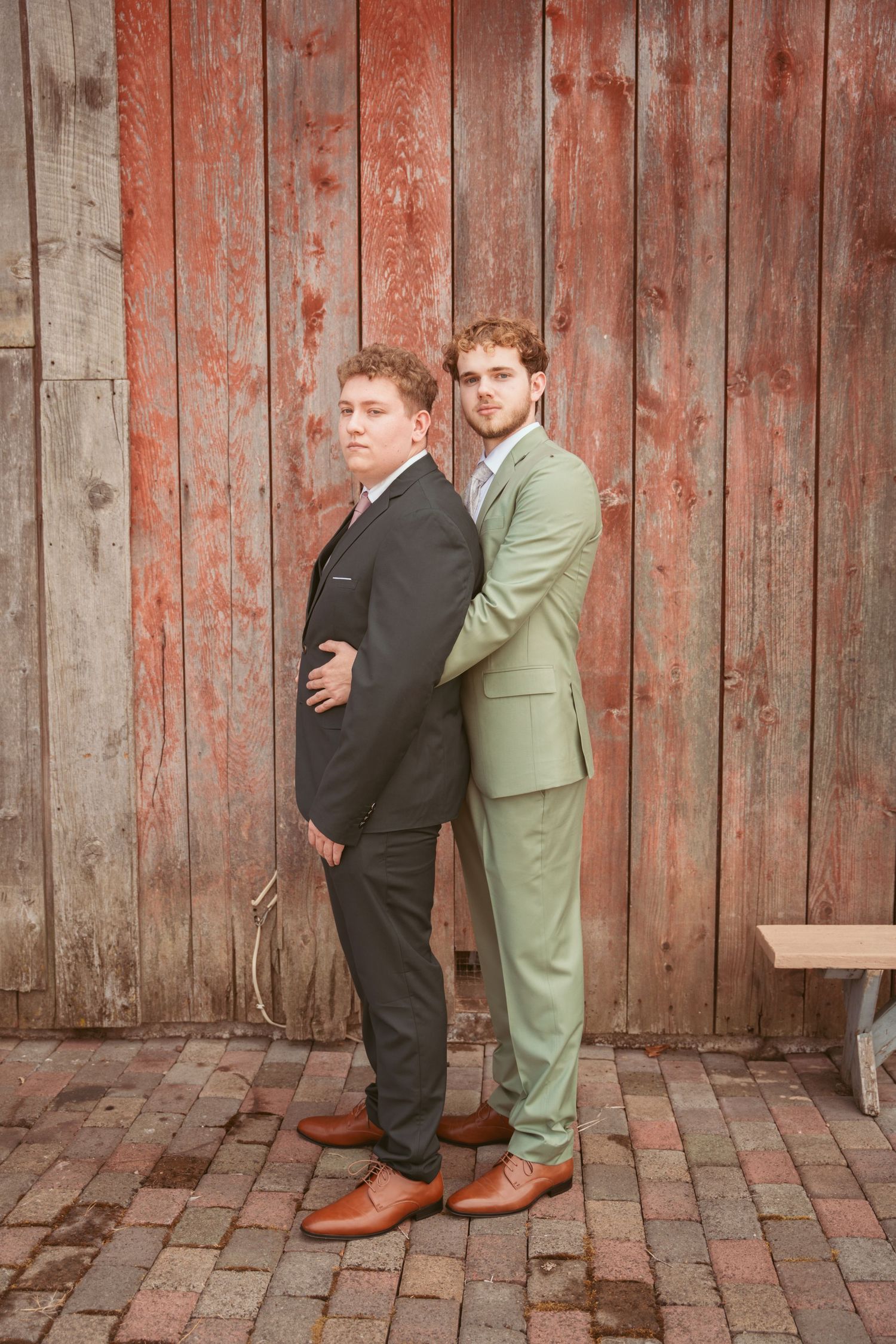 Two men in contrasting suits pose back-to-back against a rustic red barn wall.