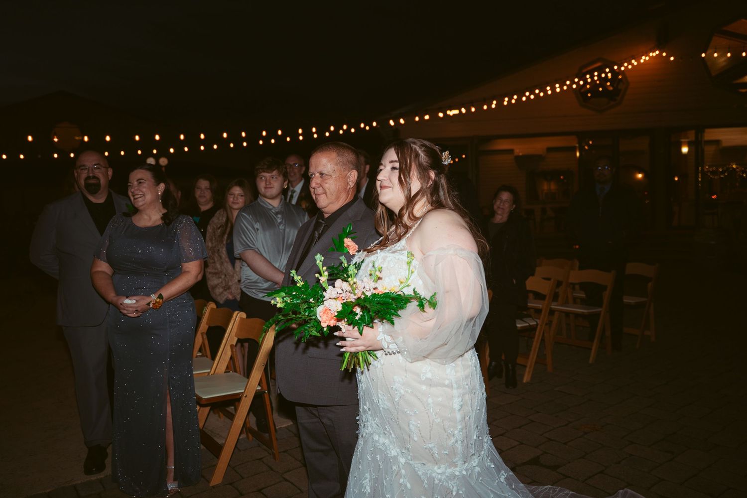 Outdoor wedding reception with string lights and wooden chairs set up on stone patio.