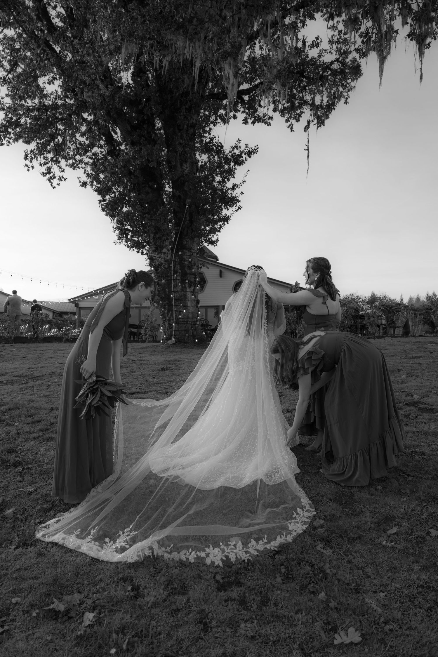 Black and white photo of bridesmaids adjusting bride's long lace veil outdoors.