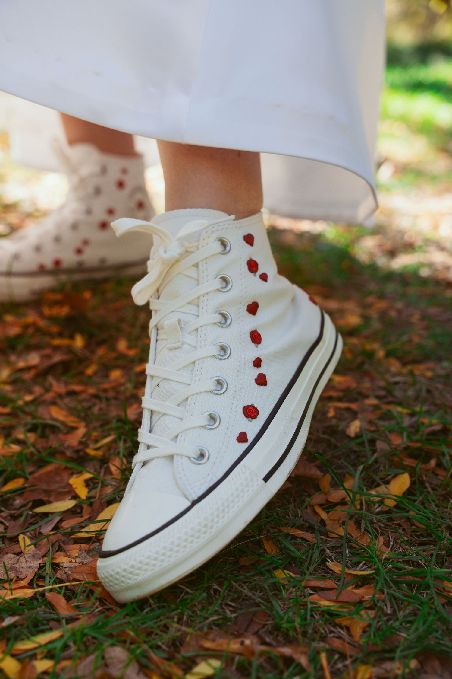 White Converse sneakers with red heart details on grass and fallen leaves.