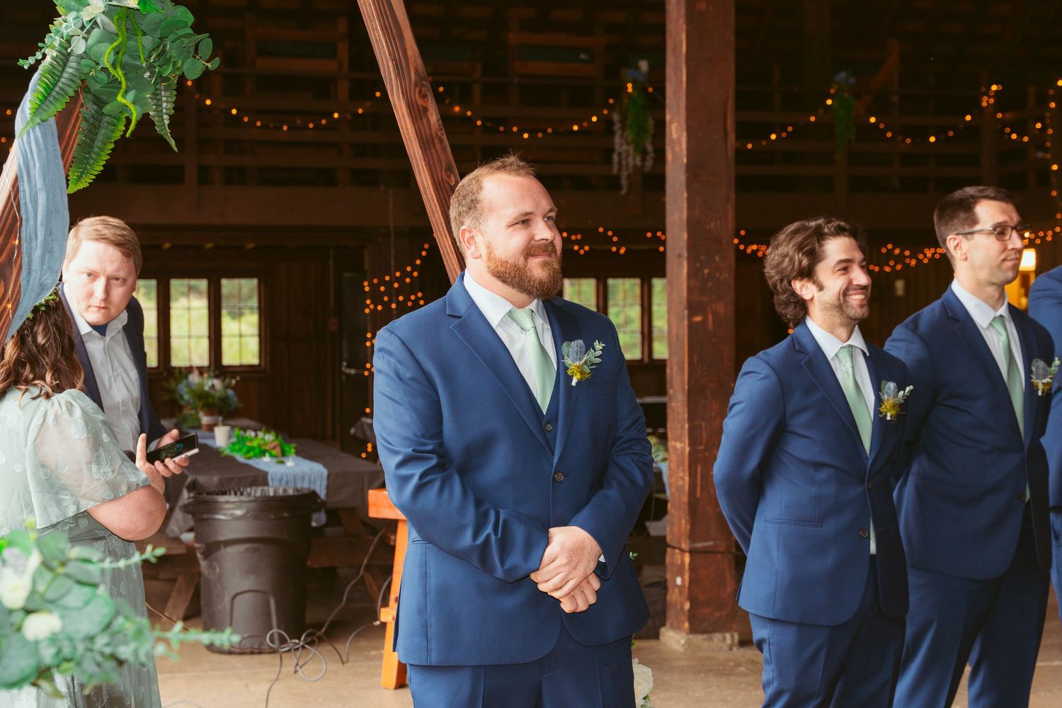 Wedding party in navy blue suits and sage green dresses stand in a rustic barn venue.